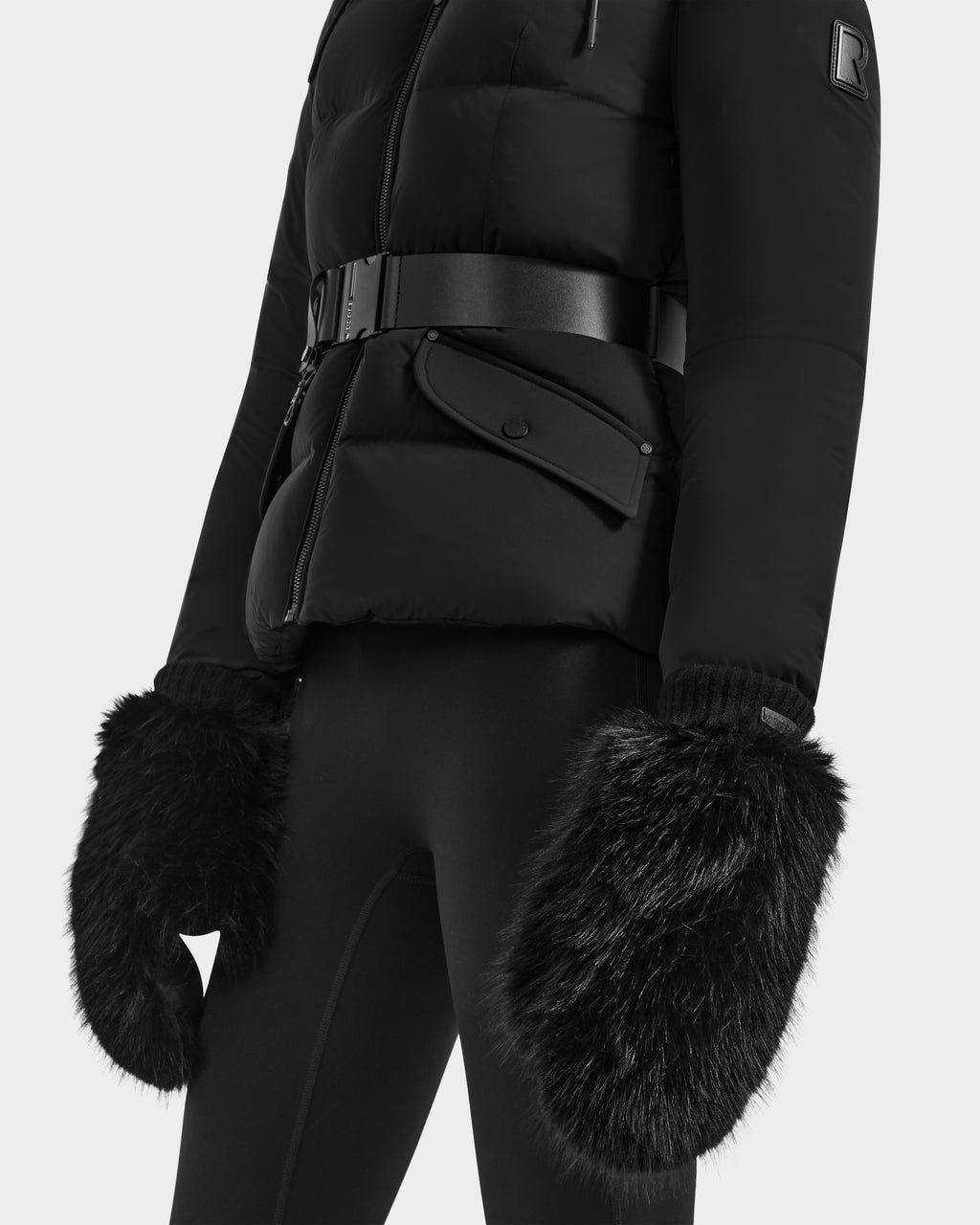A model wears the Rudsak BELLA - 8425006 Black puffer jacket with a belt, paired with black pants and oversized faux fur mittens, shown from shoulders to mid-thigh against a plain background.