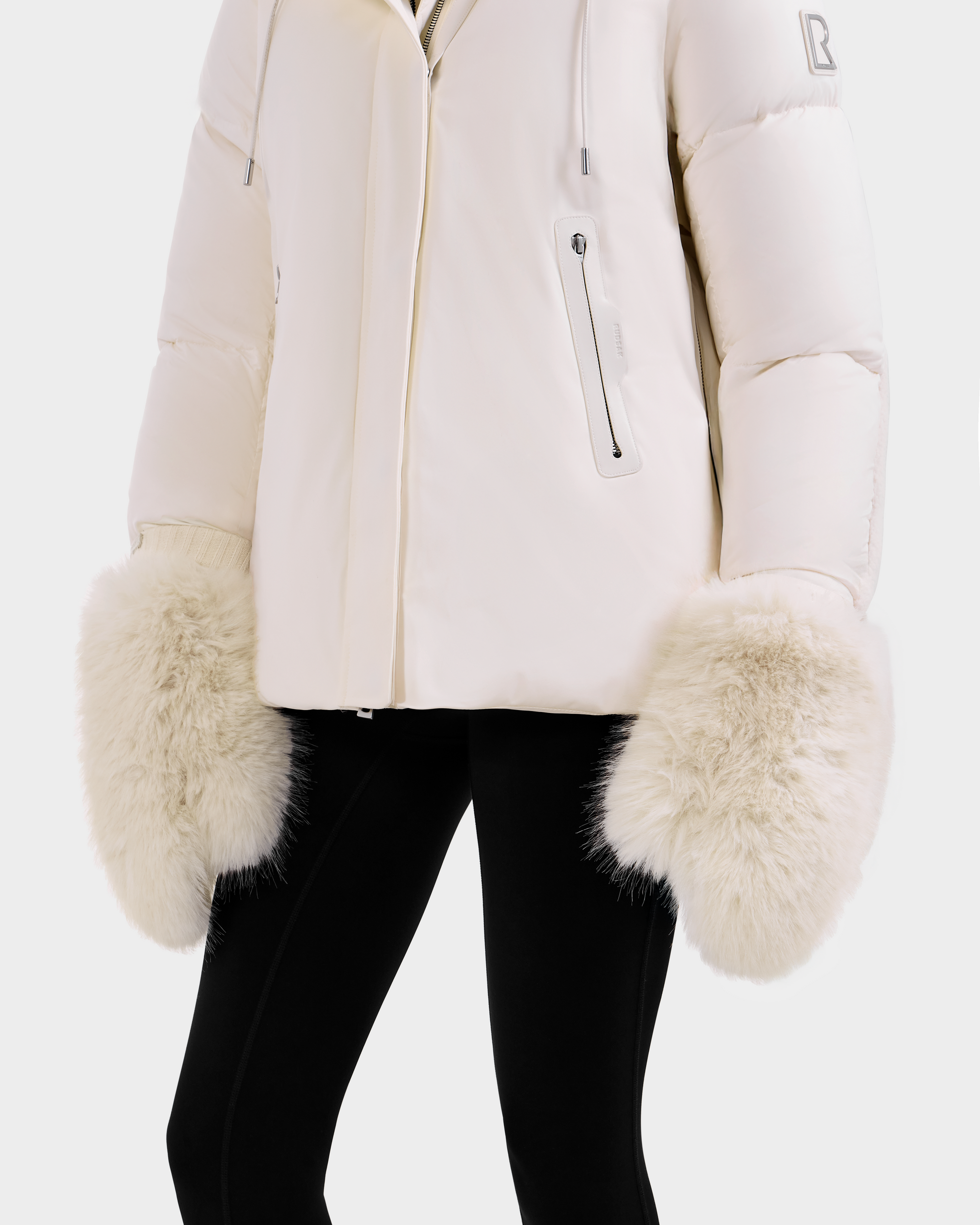 A person models the Rudsak BELLA - 8425006 Vanilla puffer jacket with large faux fur mittens and black pants, highlighting these cozy winter accessories against a plain background.
