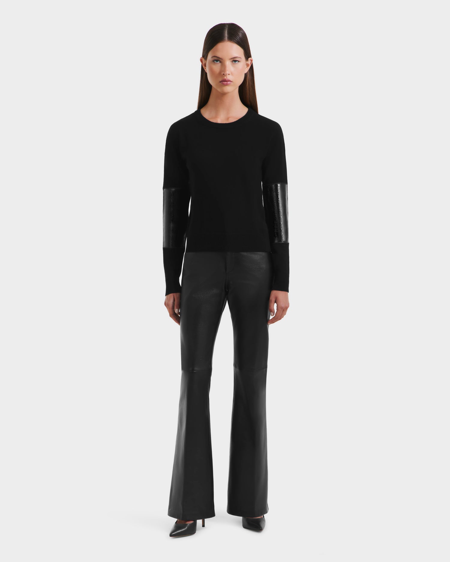 A woman faces forward in a sleek black look, wearing the Rudsak BELLEROSE - 8525554 sweater with glossy black arm patches, paired with black leather flared pants and heels against a plain white background.