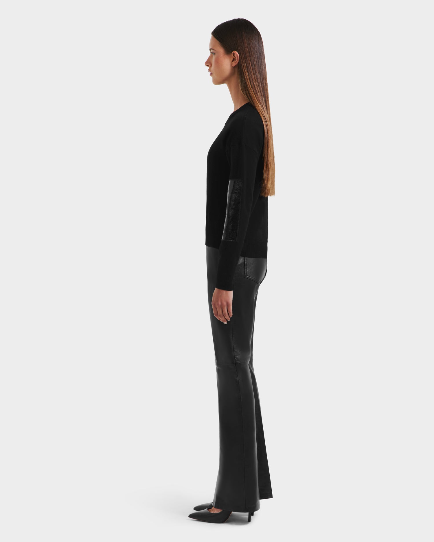 A woman with long straight brown hair stands in profile, wearing the Rudsak BELLEROSE - 8525554 Black sweater with leather elbow patches, black leather pants, and black shoes against a plain white background.