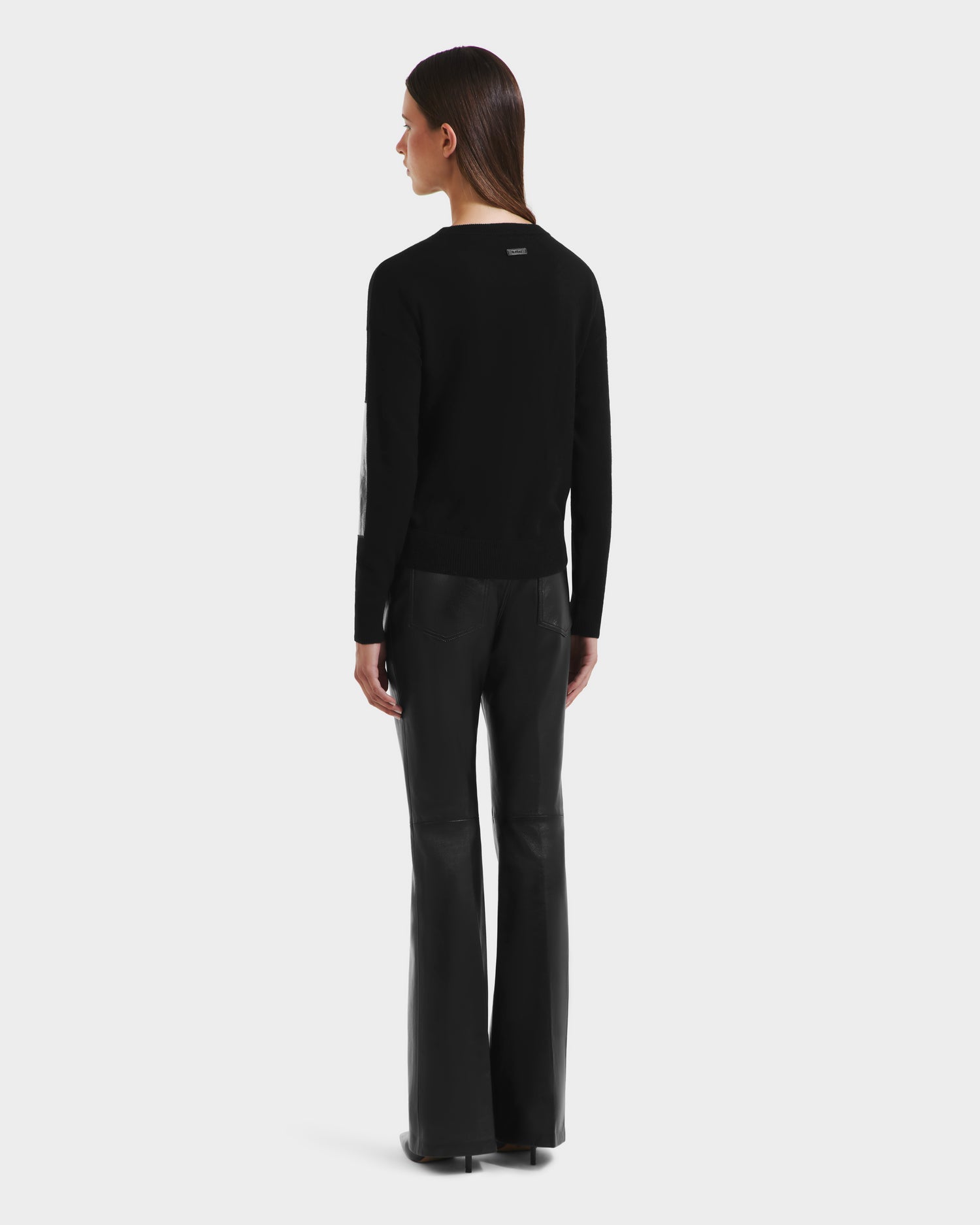 A person with straight brown hair stands facing away in a chic Rudsak BELLEROSE - 8525554 Black sweater and black leather flare pants against a plain white background.