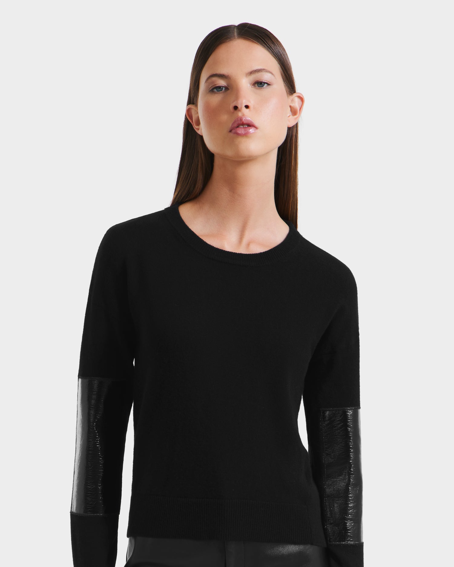 A woman with straight brown hair wears the Rudsak BELLEROSE - 8525554 Black sweater, a premium wool-blend women’s crewneck featuring glossy black textured lower sleeve panels, set against a plain light background.