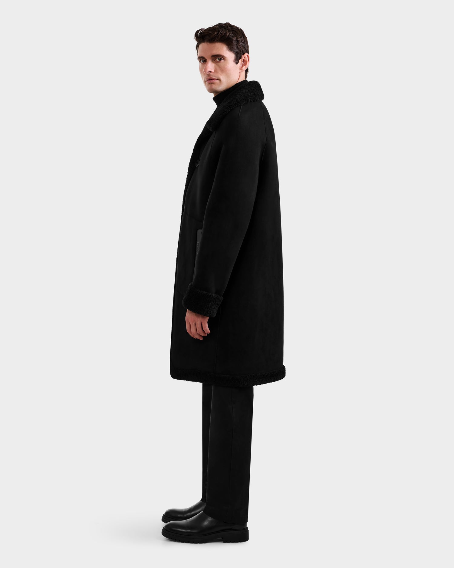 A man stands in profile against a plain light gray background, wearing Rudsak's BENITO - M125533 Black faux shearling coat, paired with black pants and black boots.