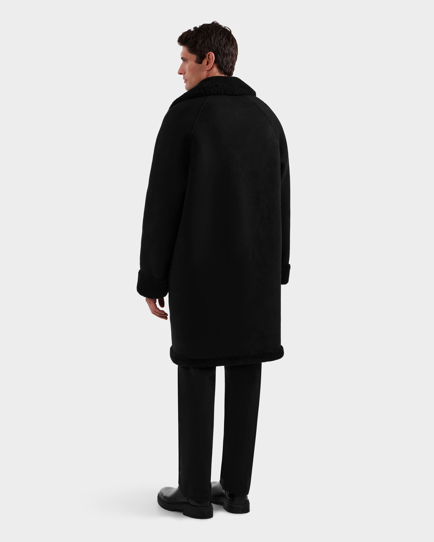 A man, seen from behind, wears the Rudsak BENITO - M125533 Black mid-length overcoat with fur collar and cuffs, paired with black pants and shoes, set against a plain light gray backdrop.
