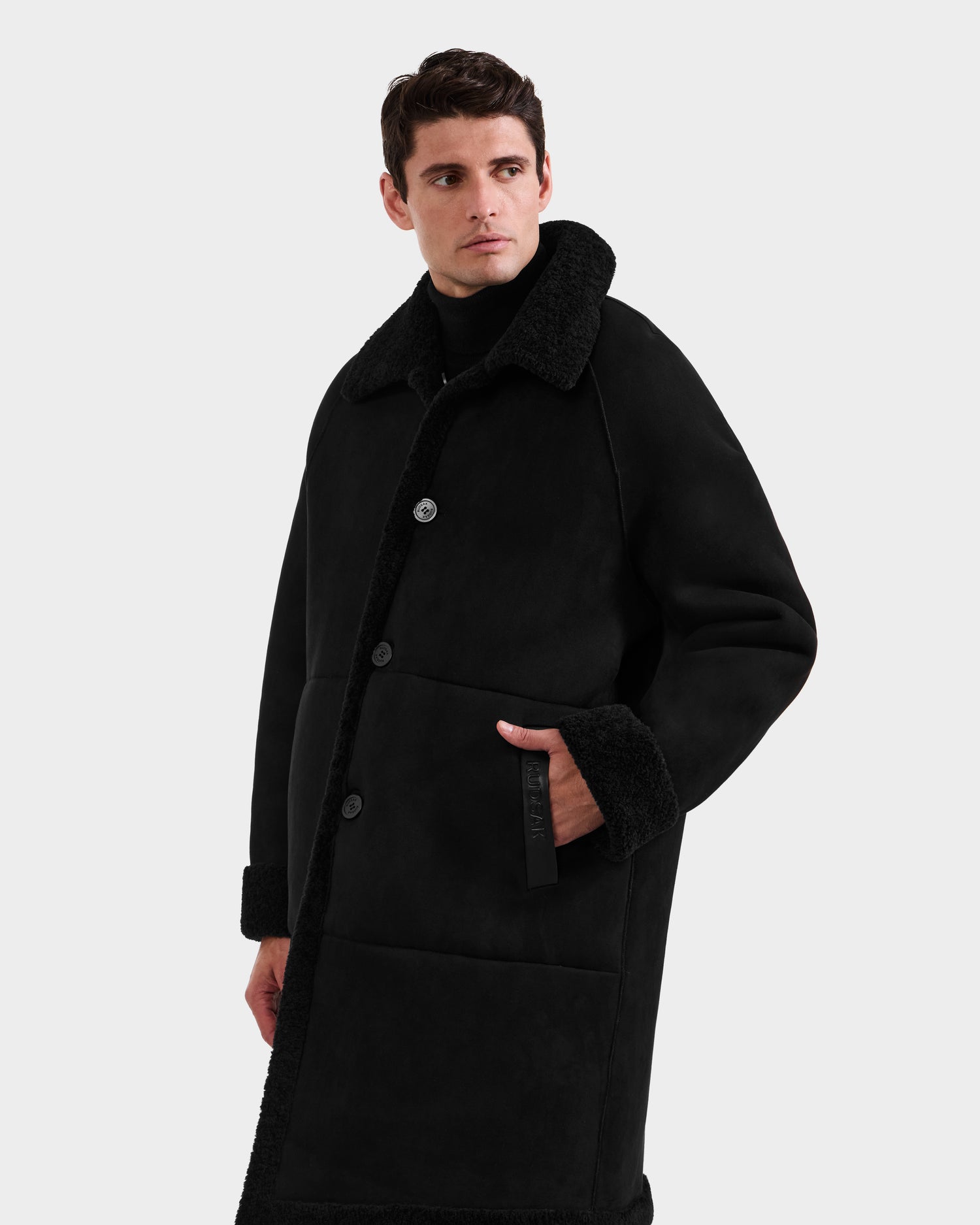 A man wears the Rudsak BENITO - M125533 Black faux shearling coat, featuring a large collar and turned-up cuffs, standing against a plain white background with one hand in his pocket and looking slightly to his left.