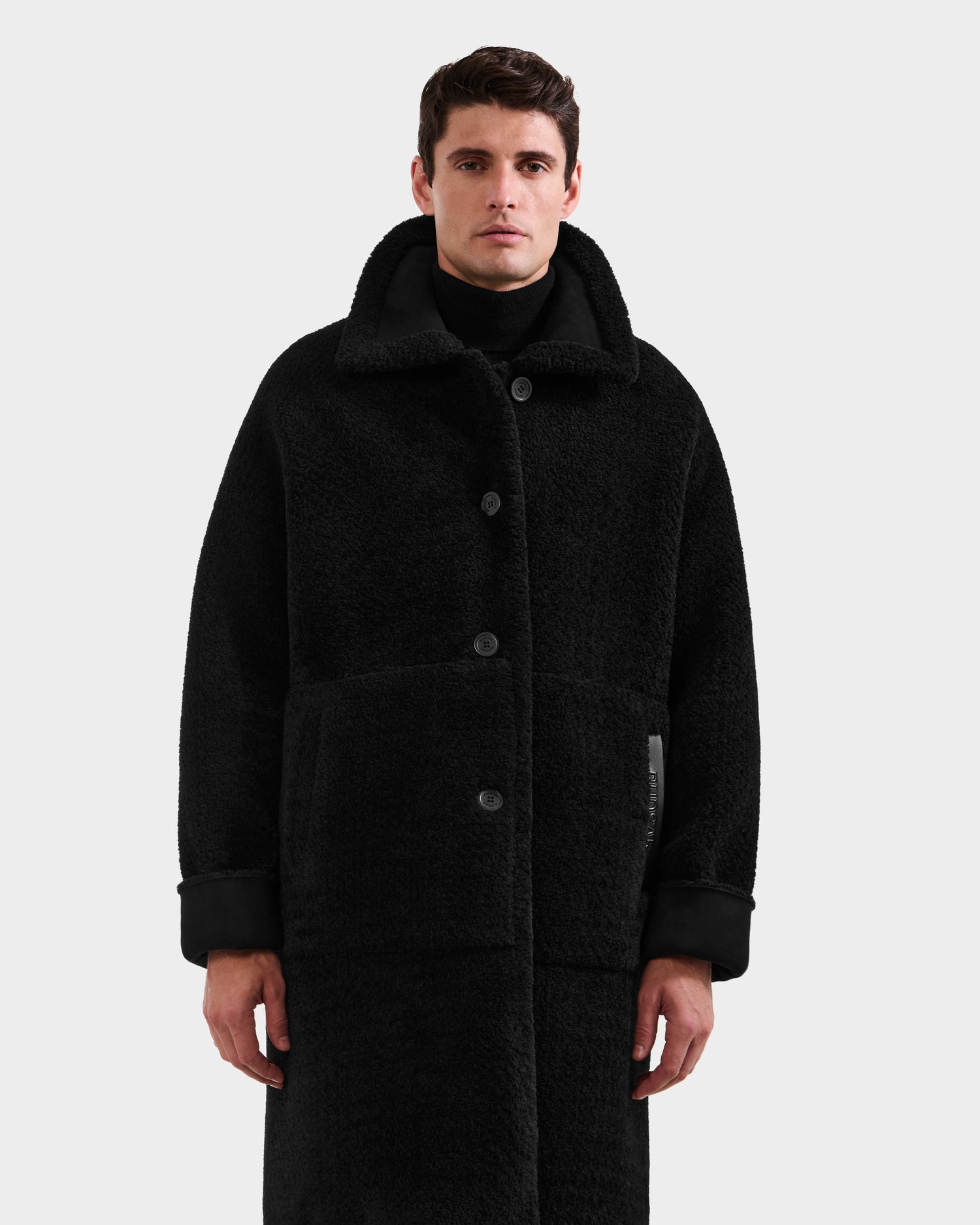 A man faces forward in a Rudsak BENITO - M125533 Black mid-length overcoat featuring large buttons, pockets, a wide collar, and textured fabric. He layers it over black attire against a plain light background.