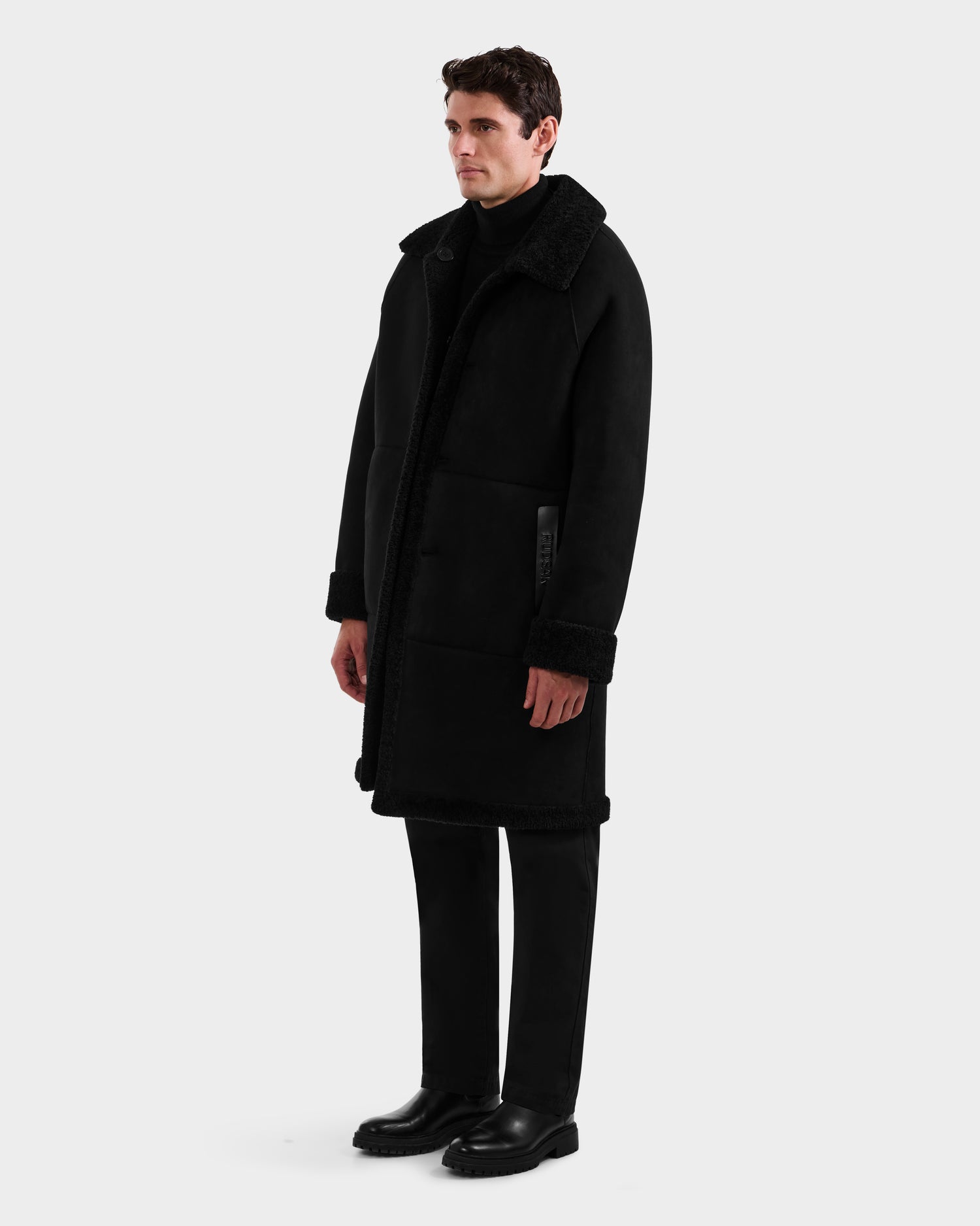 A man stands against a plain background wearing the Rudsak BENITO - M125533 Black faux shearling coat, paired with black pants, a black turtleneck, and black shoes. He is facing slightly to the side with his arms relaxed at his sides.