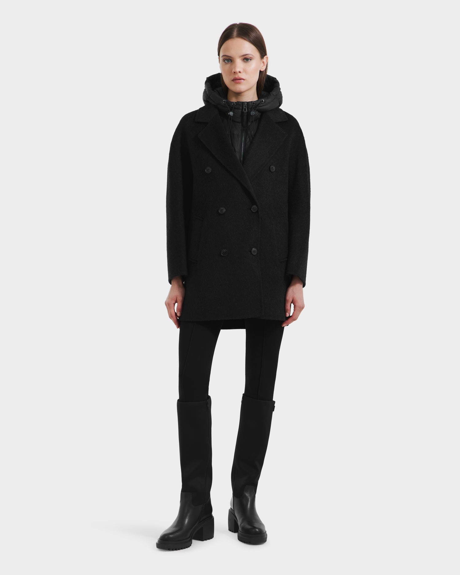 A woman faces forward against a light gray background, wearing the Rudsak BETHANY - 8125625 Black wool-silk-yak 3-in-1 coat over a black zip-up top, black pants, and knee-high boots.