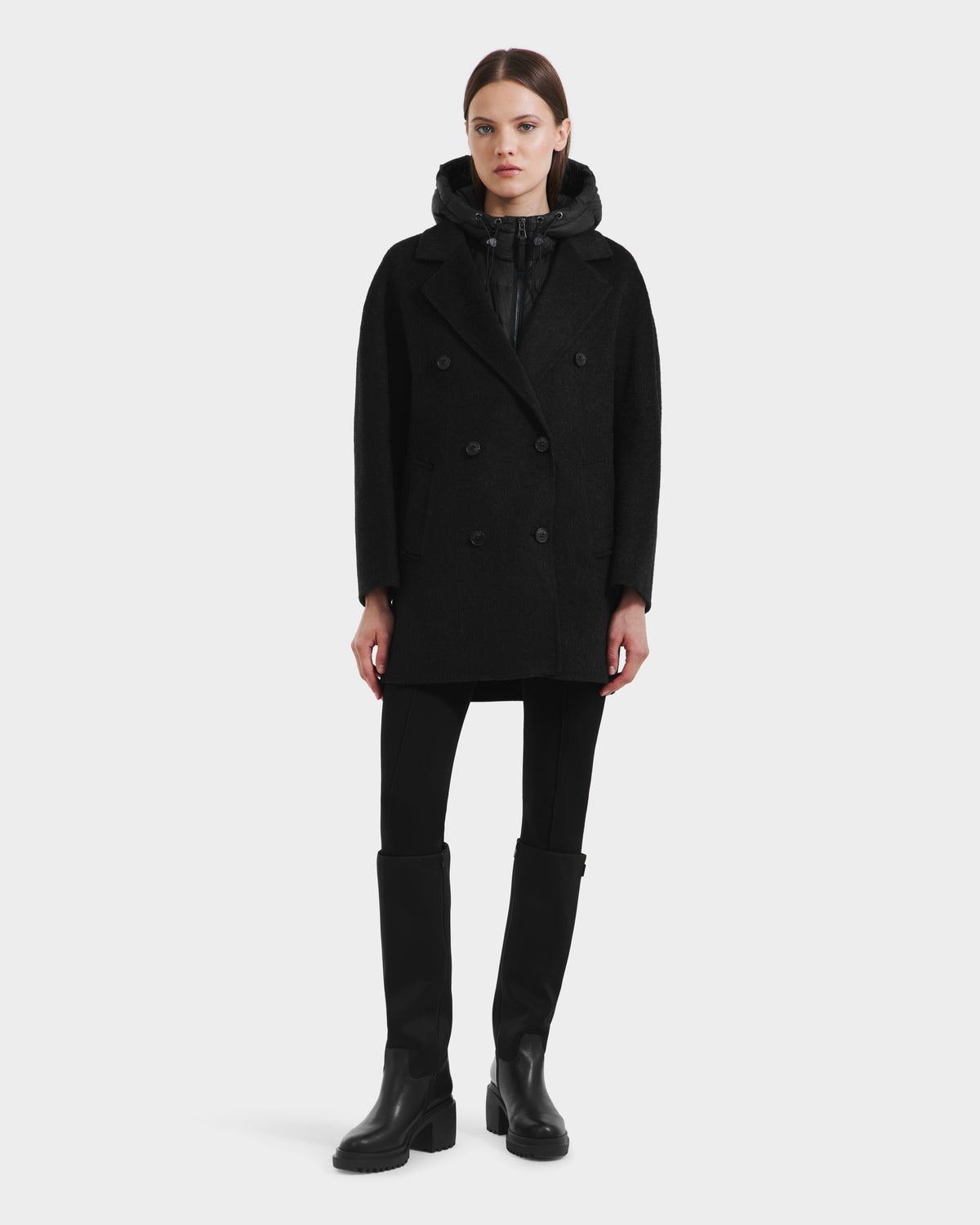 A woman faces forward against a light gray background, wearing the Rudsak BETHANY - 8125625 Black wool-silk-yak 3-in-1 coat over a black zip-up top, black pants, and knee-high boots.