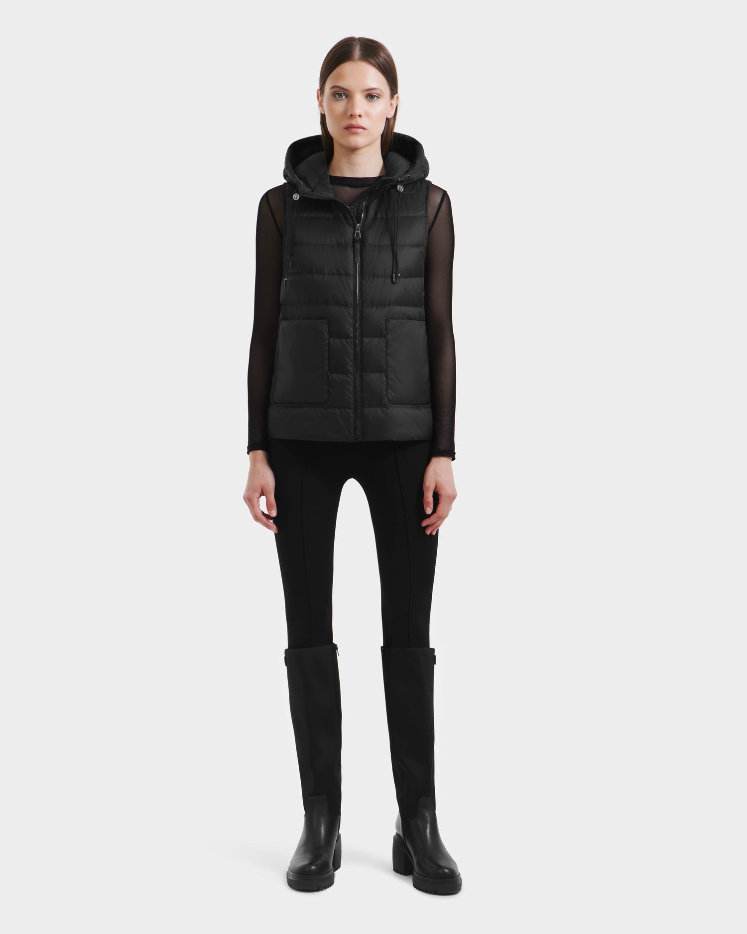 A woman faces forward, modeling the Rudsak BETHANY - 8125625 Black: a modular hooded puffer vest worn over a sheer black top, paired with fitted pants and knee-high boots, against a plain light background.