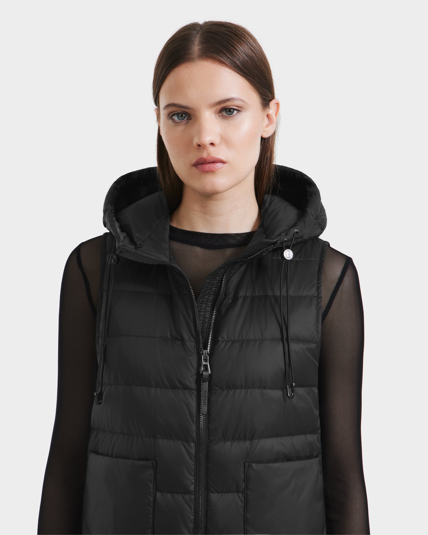 A woman with straight brown hair wears the Rudsak BETHANY - 8125625 Black hooded puffer vest over a sheer long-sleeve black top, gazing forward with a neutral expression against a plain light background.