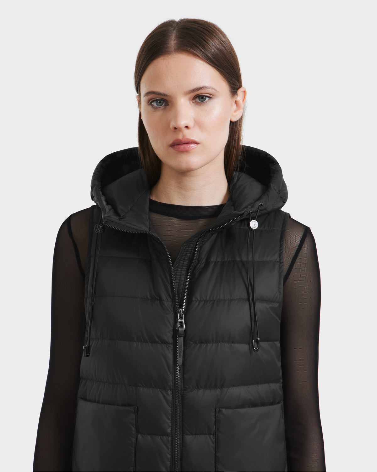 A woman with straight brown hair wears the Rudsak BETHANY - 8125625 Black hooded puffer vest over a sheer long-sleeve black top, gazing forward with a neutral expression against a plain light background.