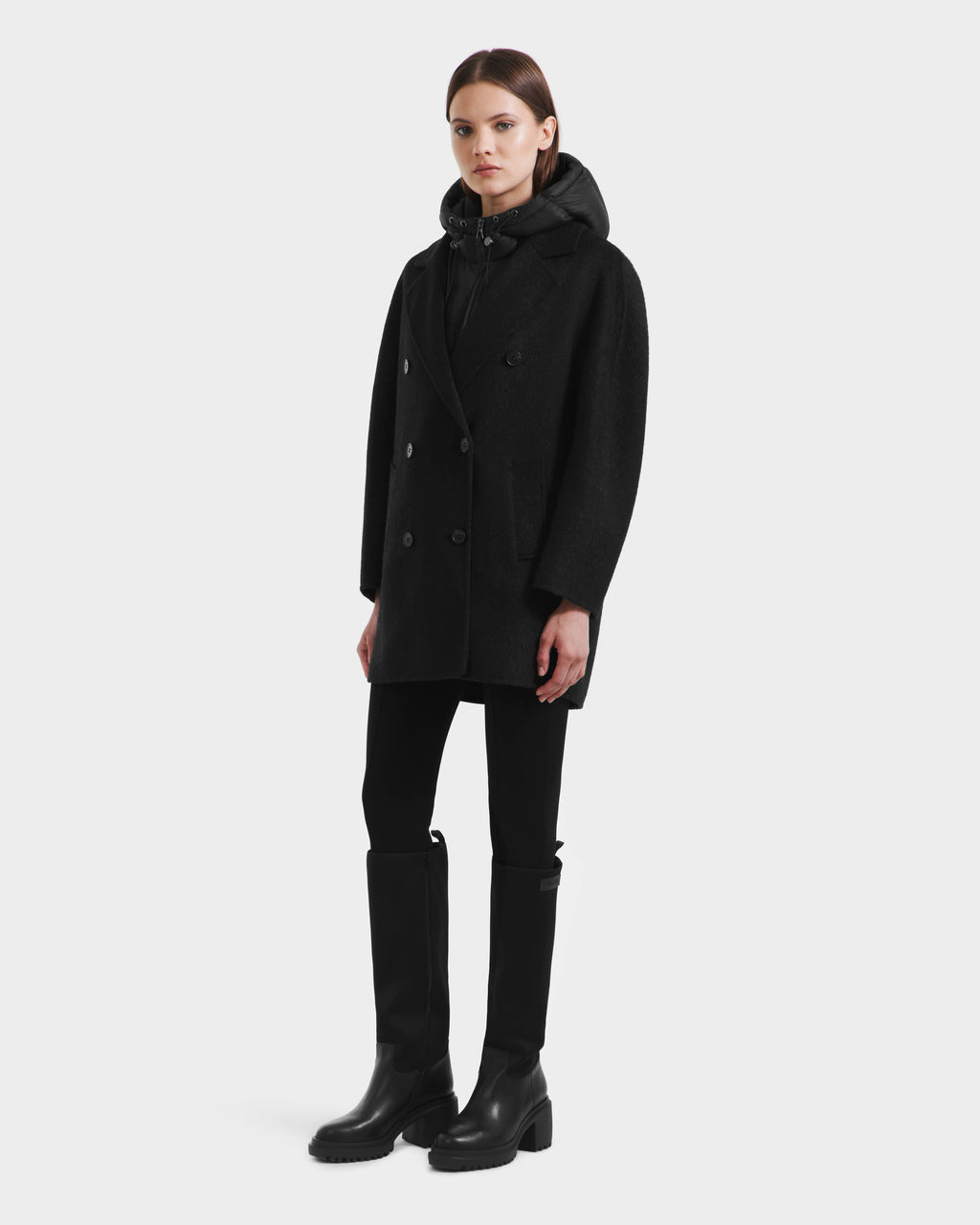 A woman faces forward in Rudsak’s BETHANY - 8125625 Black: a modular wool-silk-yak double-breasted coat layered over a zipped hoodie, black pants, and tall black boots, posed against a plain white background.
