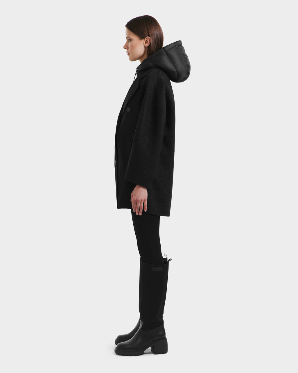A woman stands in profile wearing the Rudsak BETHANY - 8125625 Black coat with a hood, styled with black leggings and knee-high boots against a plain white background.