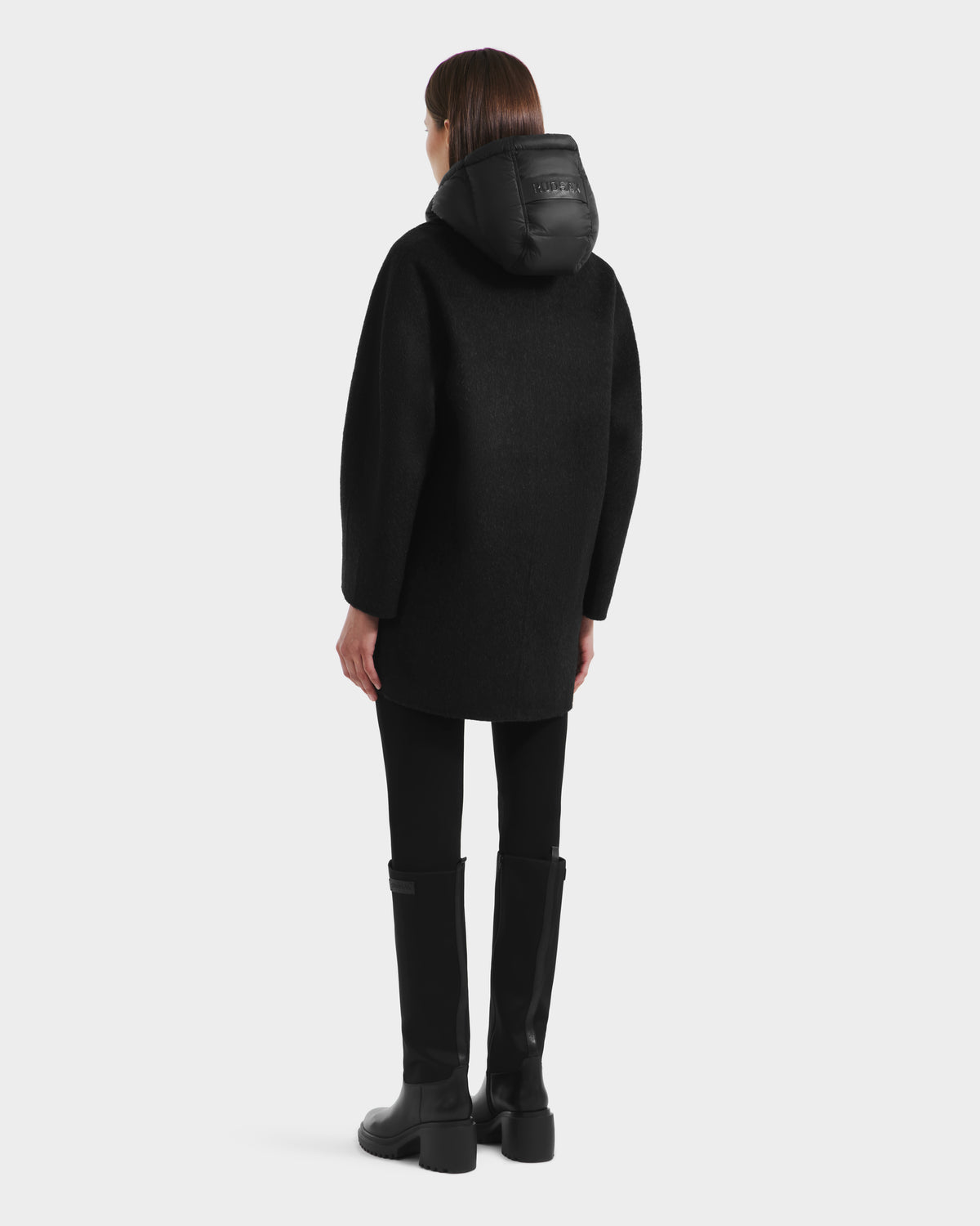 A person faces away, wearing the Rudsak BETHANY - 8125625 Black modular outerwear coat with a hood, paired with black pants and knee-high black boots against a plain light background.