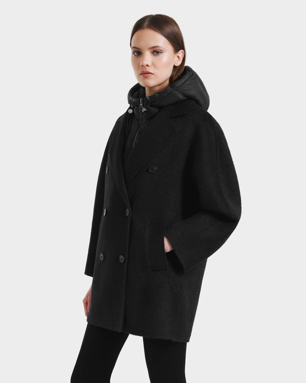 A woman wears the Rudsak BETHANY - 8125625 Black coat, featuring a 3-in-1 silhouette, over a black hoodie while standing against a light grey background and looking at the camera with a neutral expression.