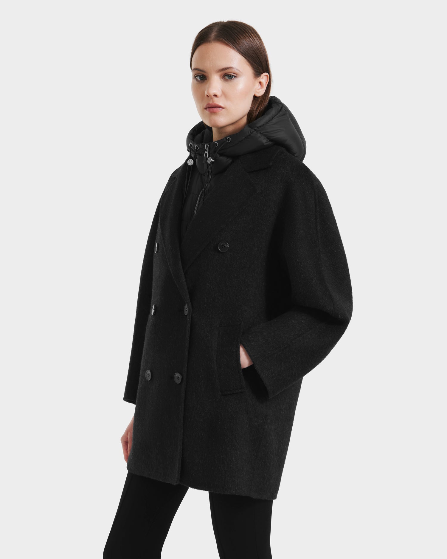 A woman wears the Rudsak BETHANY - 8125625 Black coat, featuring a 3-in-1 silhouette, over a black hoodie while standing against a light grey background and looking at the camera with a neutral expression.