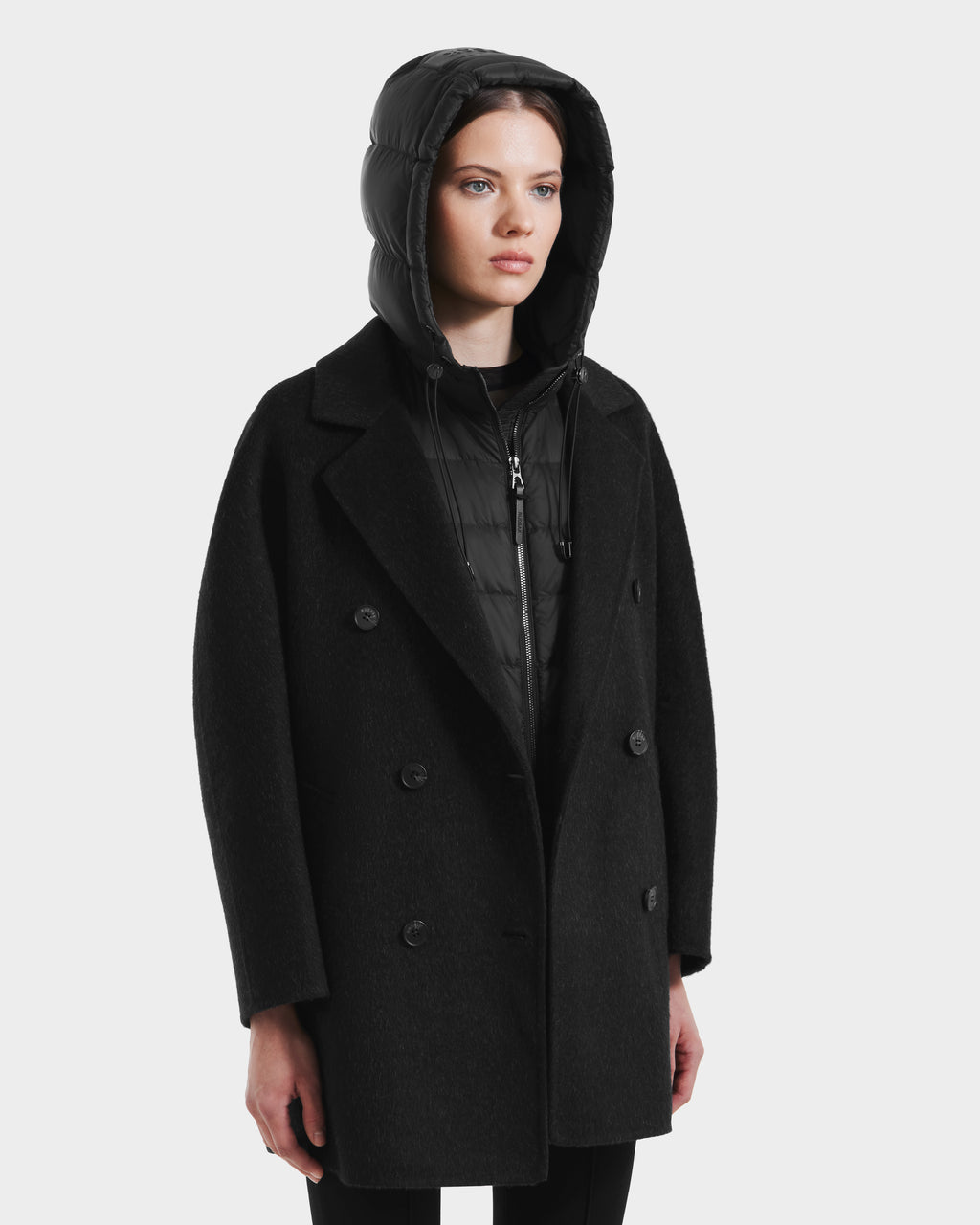 A woman wears the Rudsak BETHANY - 8125625 Black modular outerwear—a double-breasted coat layered over a zip-up puffer jacket with the hood up—standing against a plain white background.