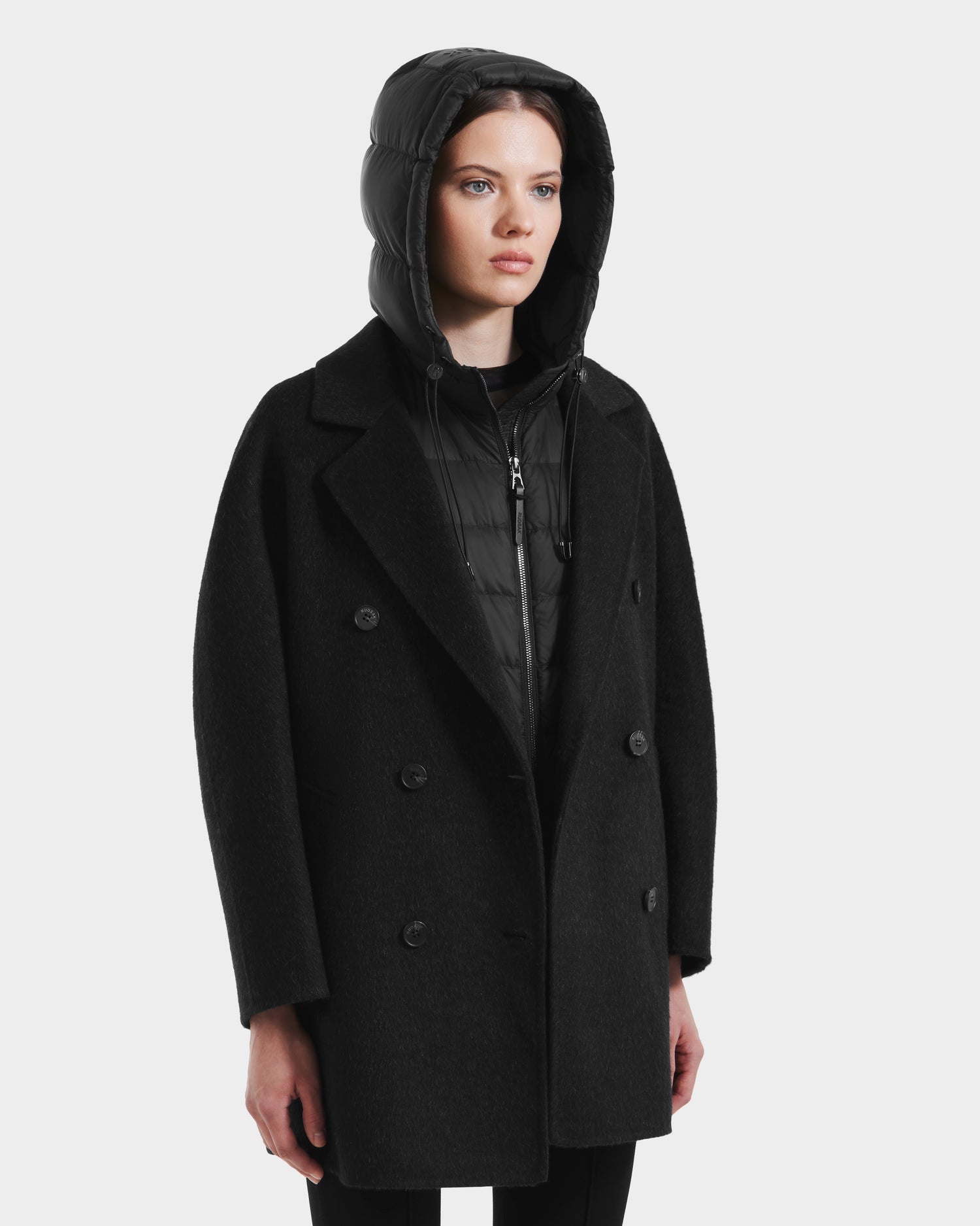 A woman wears the Rudsak BETHANY - 8125625 Black modular outerwear—a double-breasted coat layered over a zip-up puffer jacket with the hood up—standing against a plain white background.