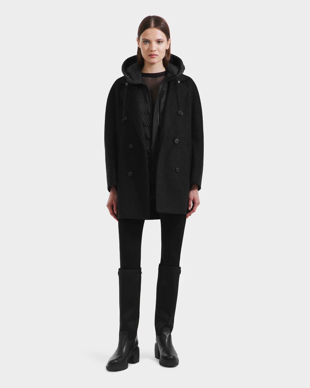 A woman faces forward against a white background, wearing the Rudsak BETHANY - 8125625 Black modular hooded coat over a zip-up jacket, with black pants and knee-high boots.