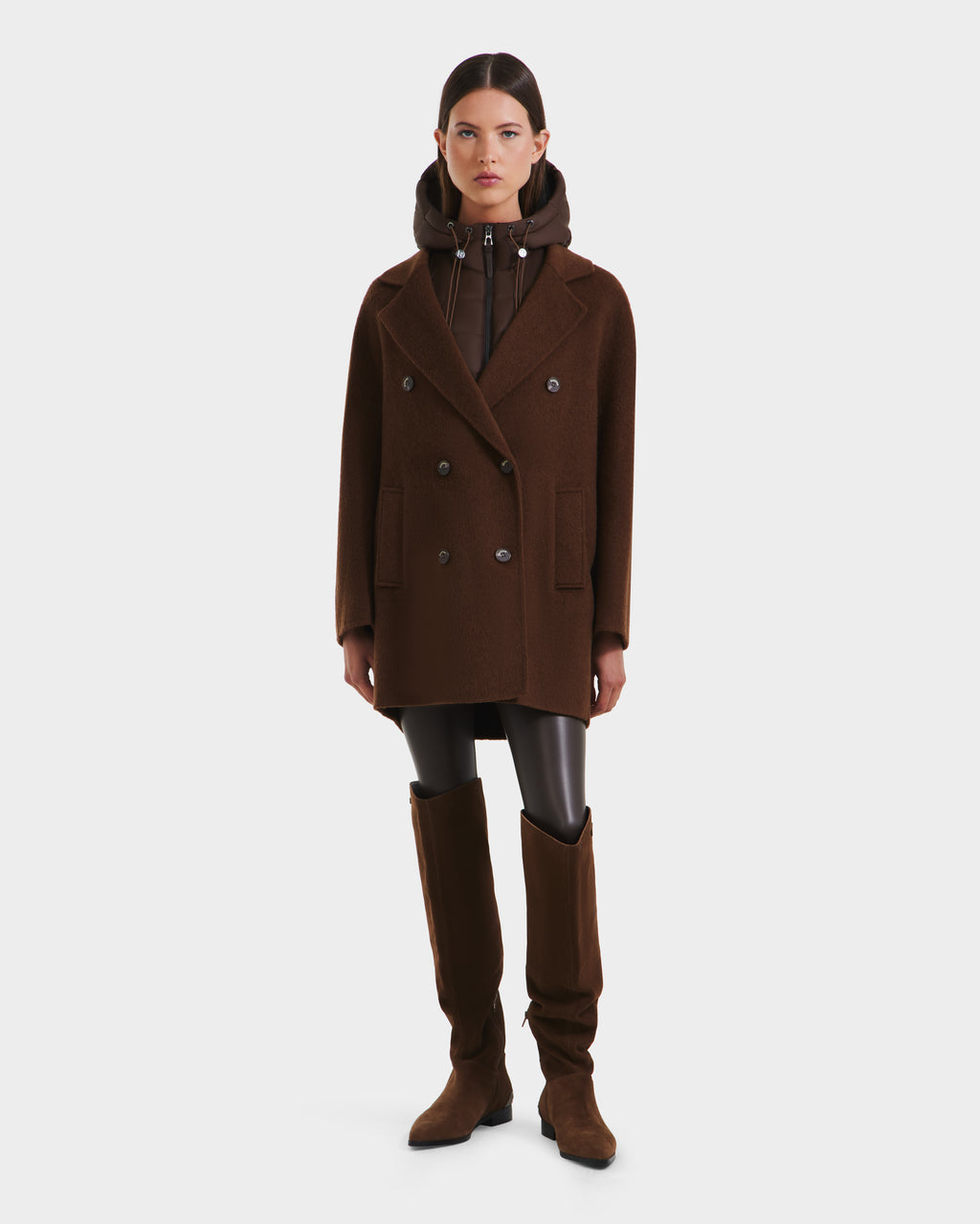 A woman stands against a plain background wearing Rudsak’s BETHANY - 8125625 Mocha double-breasted coat over a hooded jacket, paired with black leggings and knee-high brown boots.