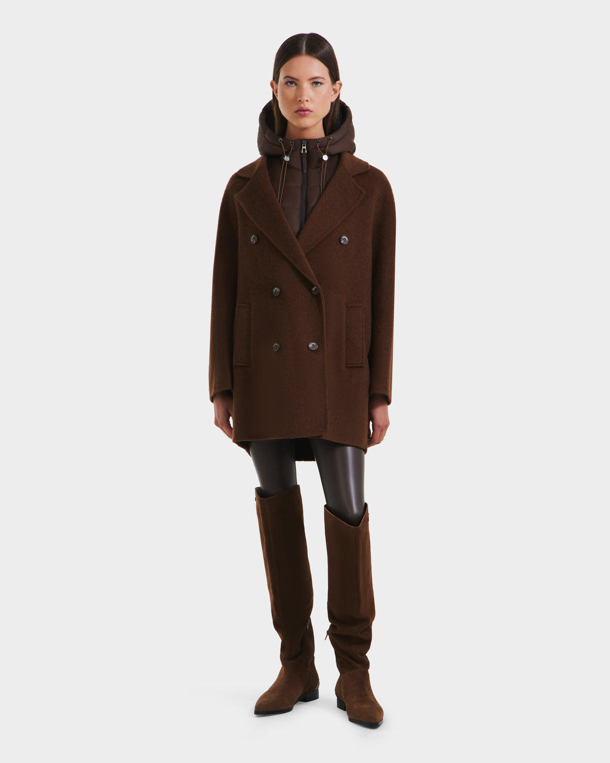 A woman stands against a plain background wearing Rudsak’s BETHANY - 8125625 Mocha double-breasted coat over a hooded jacket, paired with black leggings and knee-high brown boots.