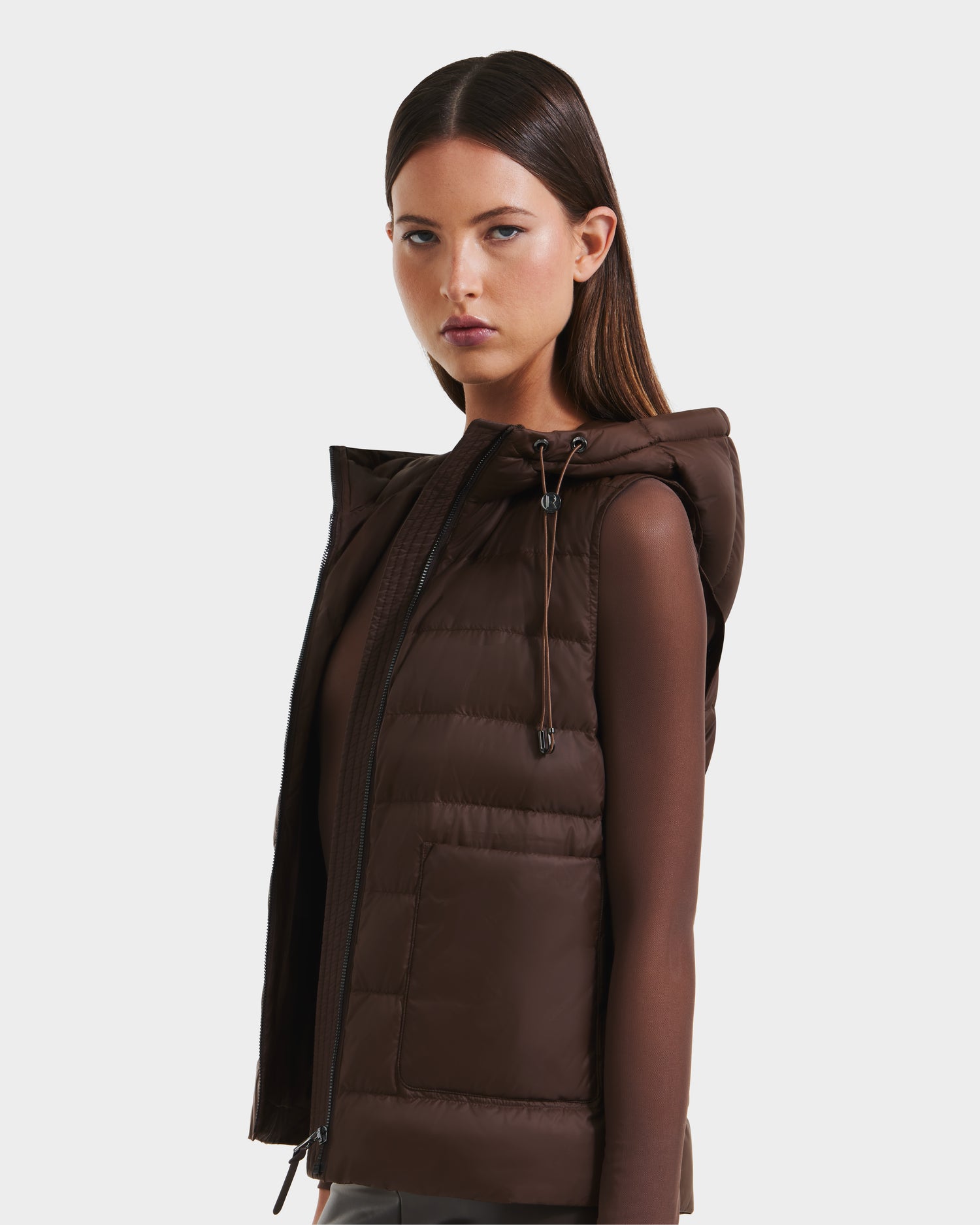 A woman with straight brown hair models the Rudsak BETHANY - 8125625 Mocha sleeveless hooded vest over a sheer brown long-sleeve top, creating an effortlessly modular outerwear look against a plain background.