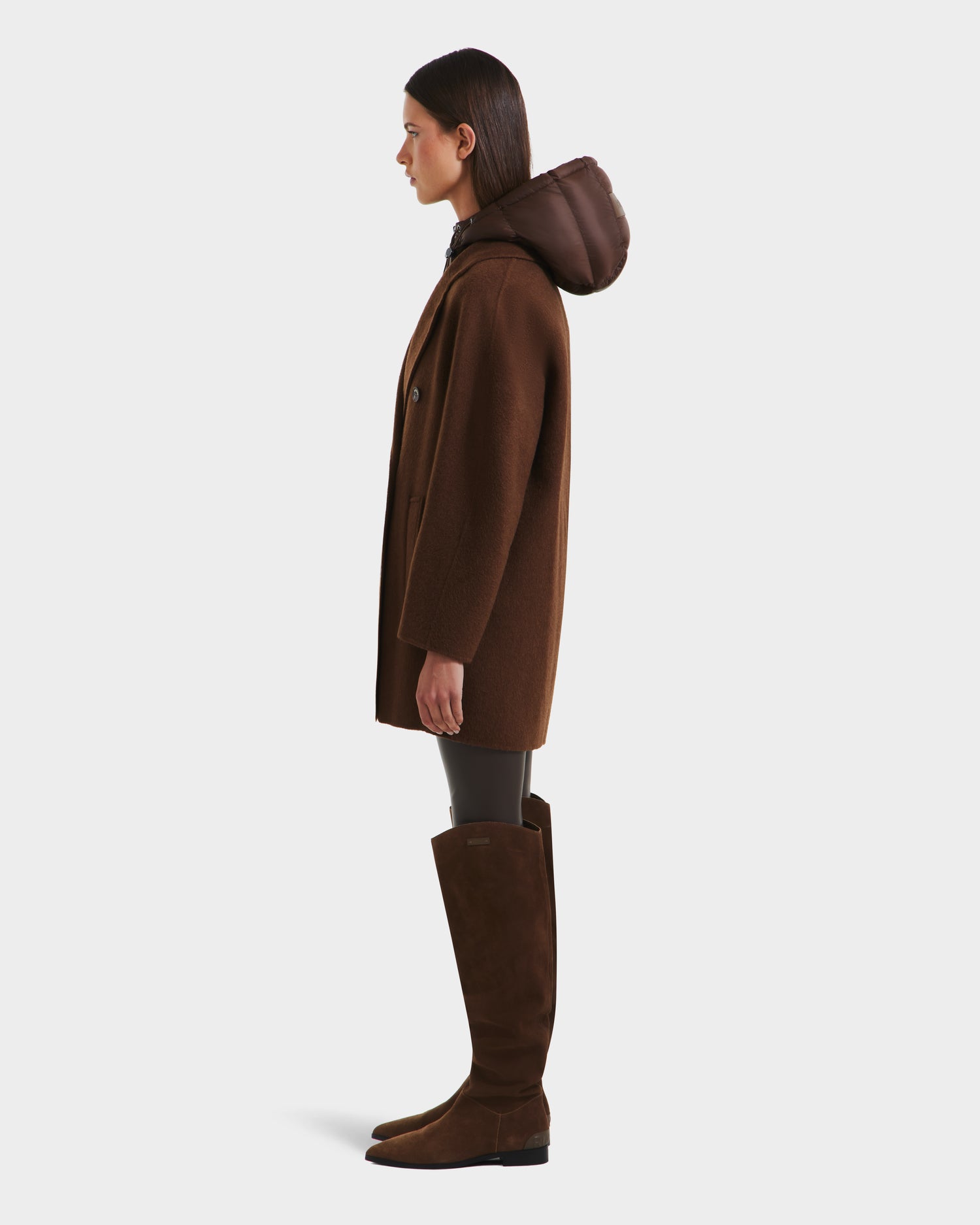 A woman stands in profile wearing the Rudsak BETHANY - 8125625 Mocha modular coat with a hood, paired with dark leggings and knee-high brown suede boots, set against a plain light background.