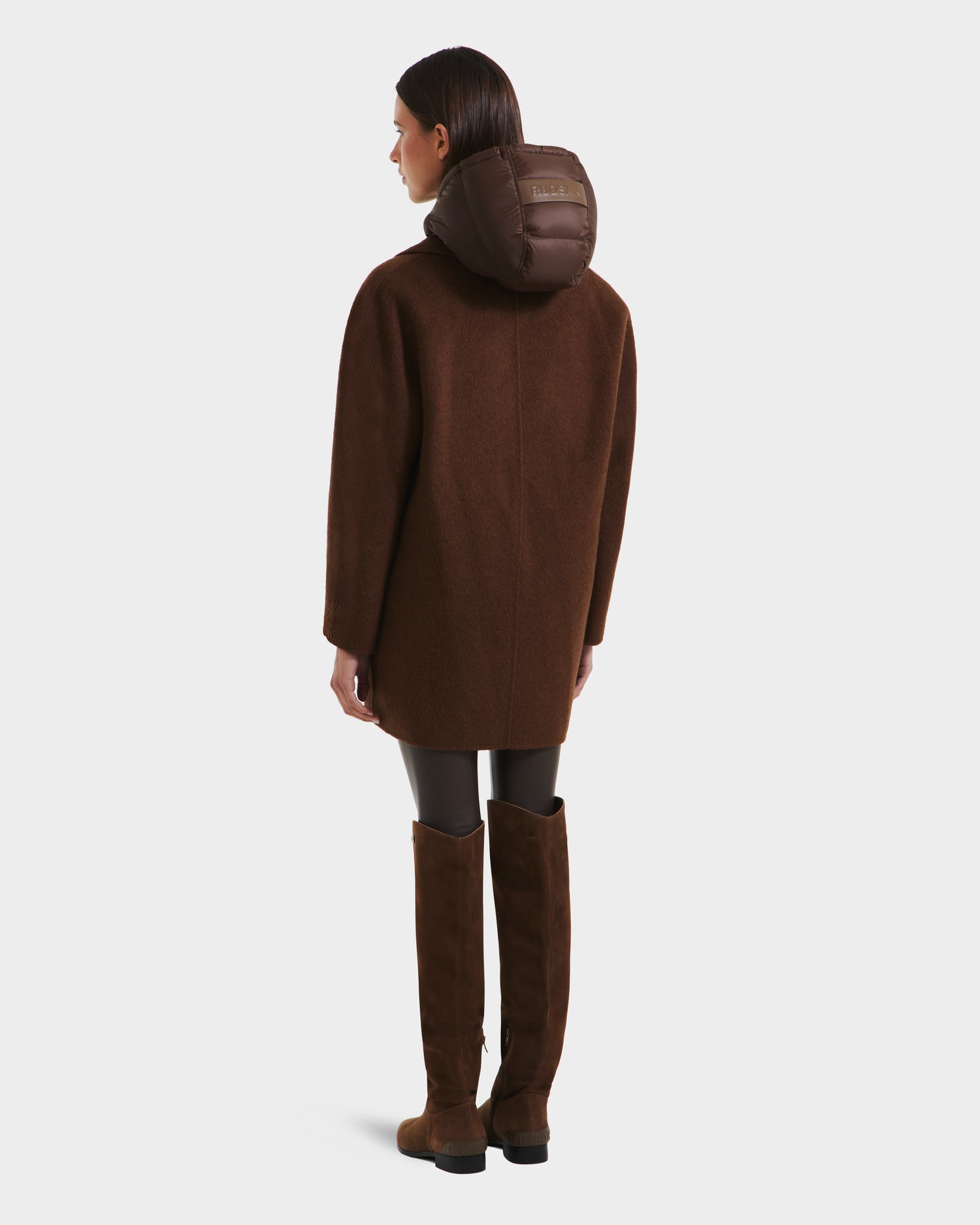 A person faces away, wearing the Rudsak BETHANY - 8125625 Mocha double-breasted coat with padded hood, paired with black leggings and knee-high brown boots against a plain white background.
