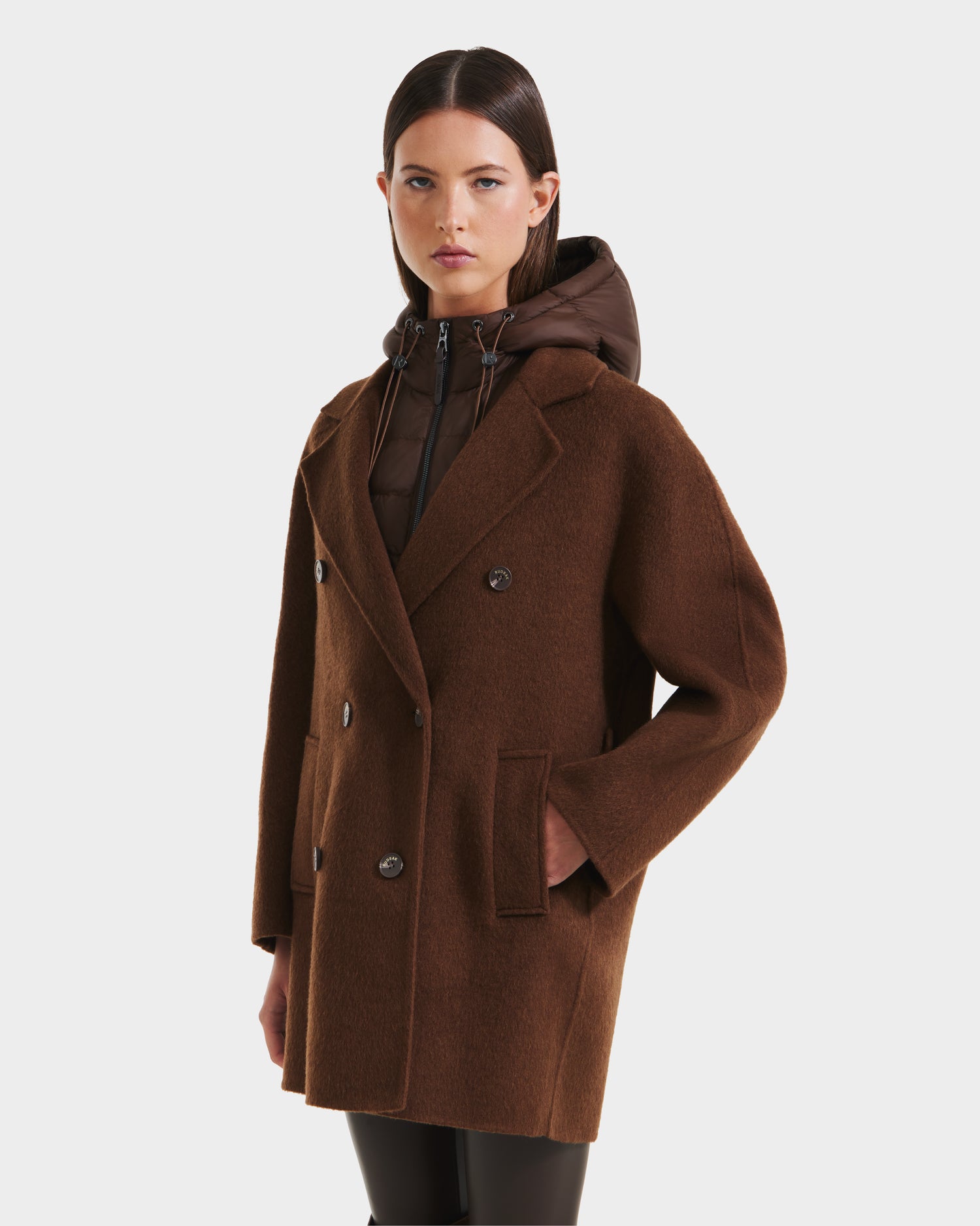 A woman wears the Rudsak BETHANY - 8125625 Mocha: a brown double-breasted coat with large lapels and side pockets over a hooded padded jacket, standing against a plain white background.
