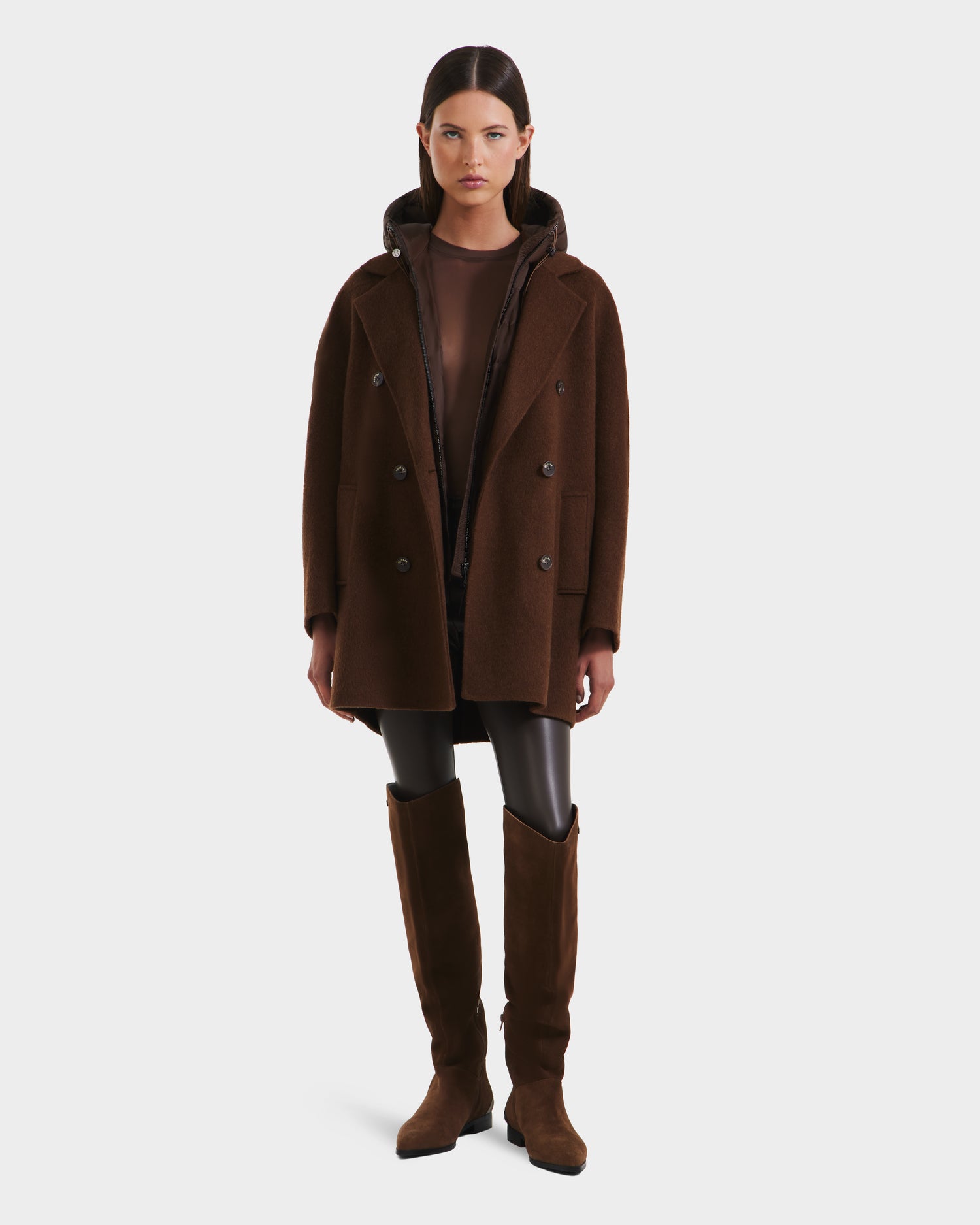 A woman stands facing forward in Rudsak’s BETHANY - 8125625 Mocha double-breasted coat with a 3-in-1 silhouette, a sheer brown top, black leggings, and knee-high brown suede boots against a plain white background.