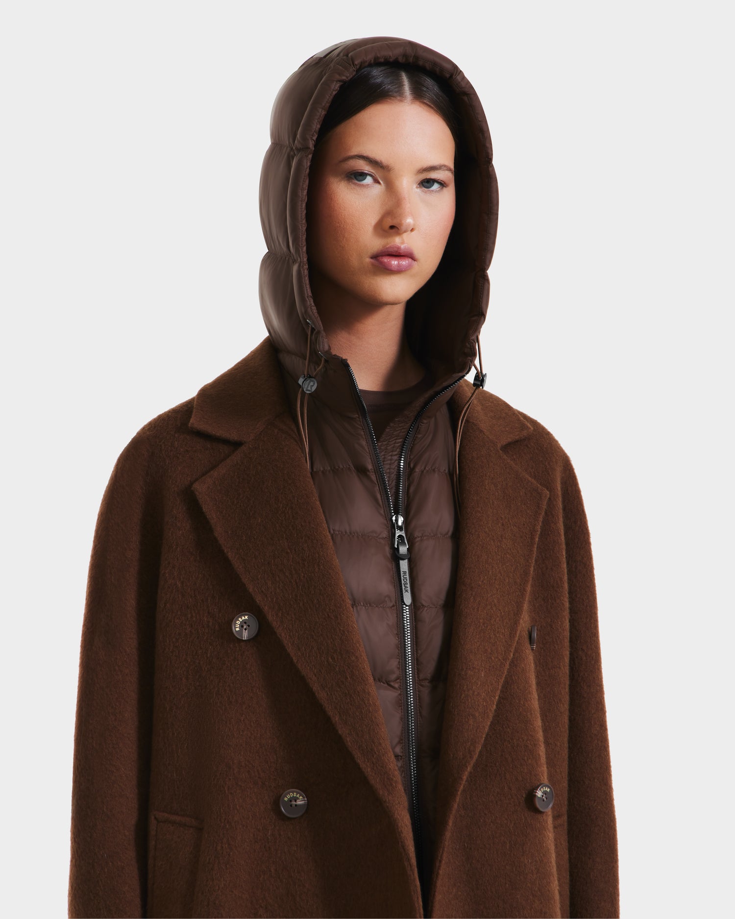 A woman models the Rudsak BETHANY - 8125625 Mocha double-breasted coat layered over a hooded puffer, highlighting versatile outerwear against a plain white background.
