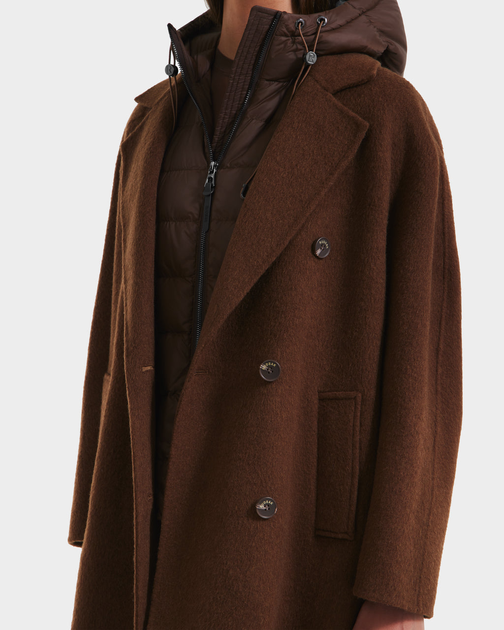 A person wears the Rudsak BETHANY - 8125625 Mocha double-breasted coat over a brown quilted puffer jacket, creating a modern 3-in-1 silhouette, shown from shoulders to mid-thigh against a white background.