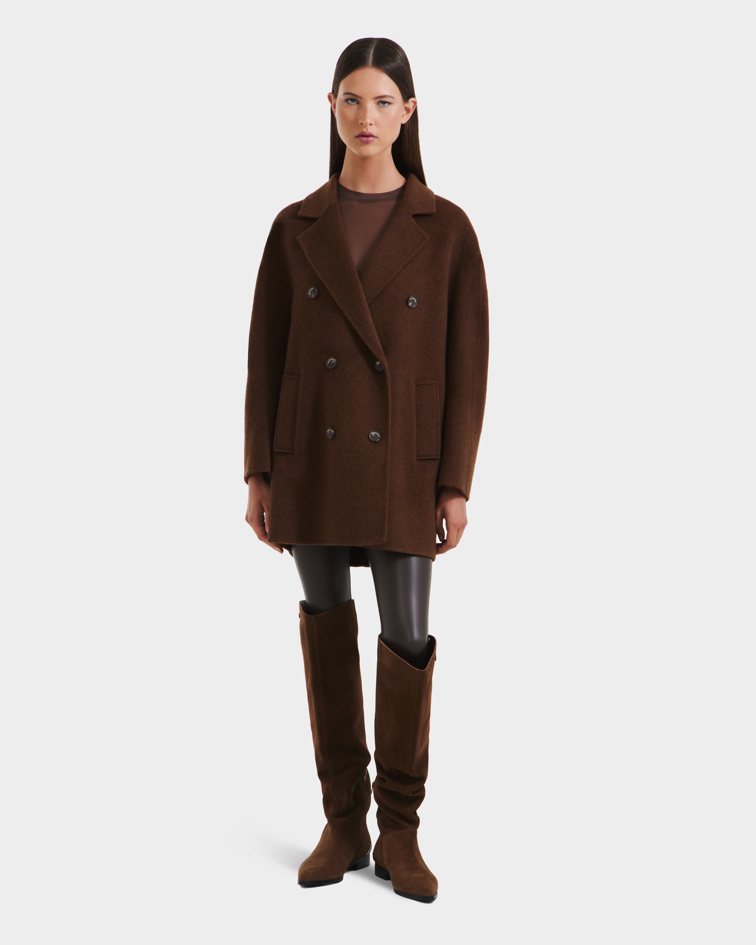 A woman with straight dark hair and a neutral expression wears the Rudsak BETHANY - 8125625 Mocha double-breasted coat over a sheer brown top, black leggings, and tall suede boots, standing against a plain white background.