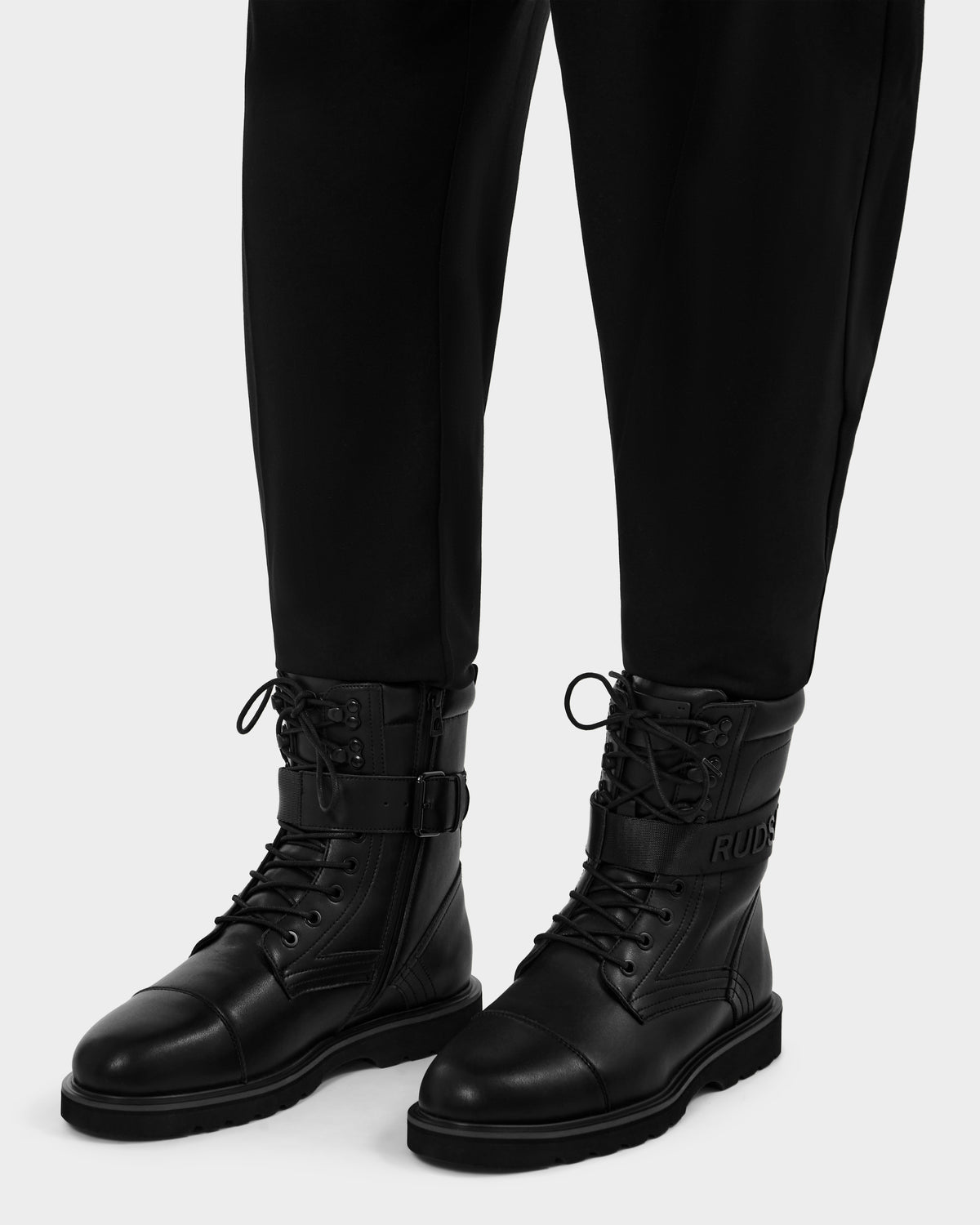 A person wears black pants and Rudsak BLITZ - M223500 Black military-inspired leather lace-up boots with side buckles and "RUDSAK" embossed on the ankle, set against a plain light background.