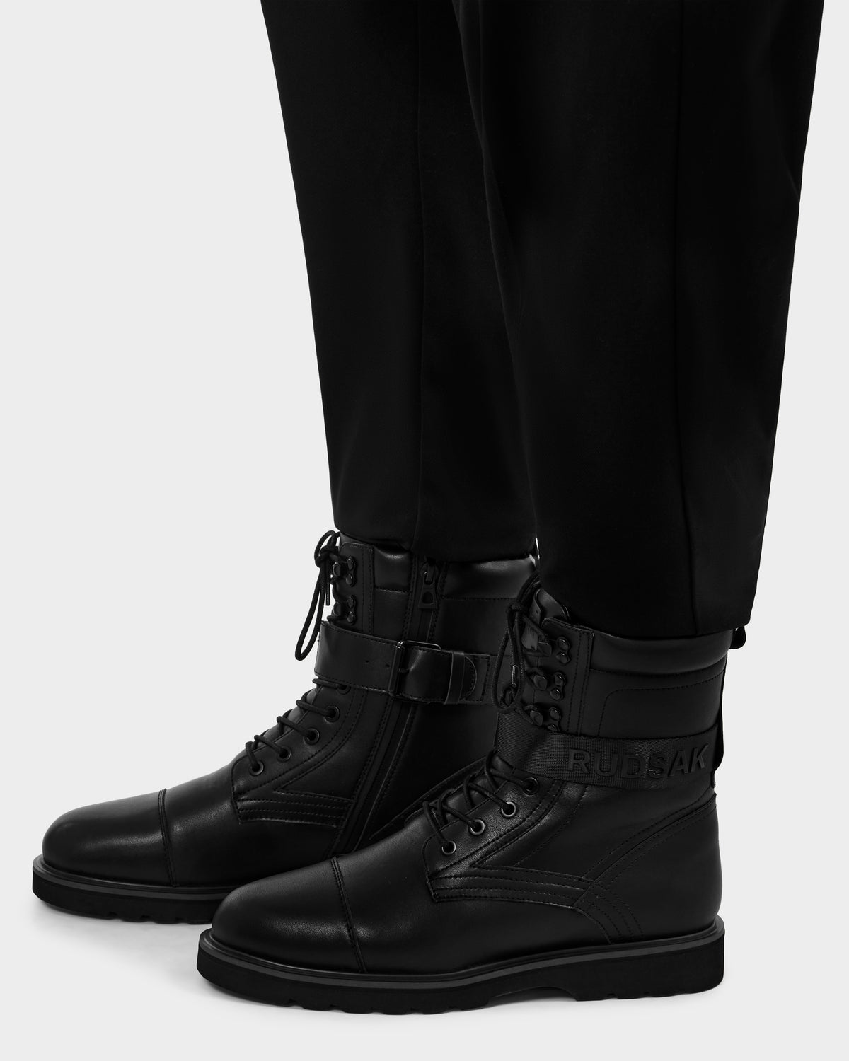 A person wears black Rudsak BLITZ - M223500 men's leather lace-up boots with zip closures, ankle strap and buckle detail, and a rugged sole for a military-inspired look, paired with black pants.