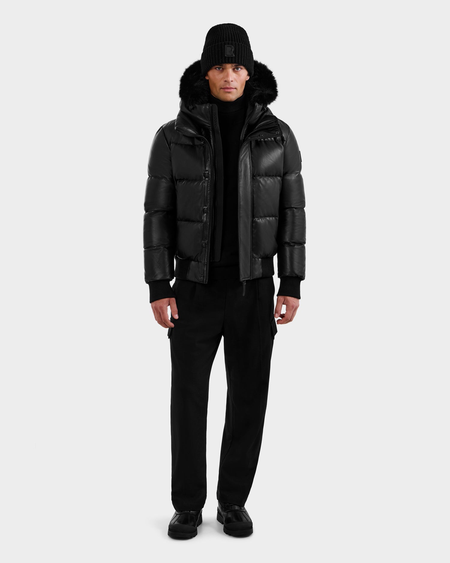 A man faces forward against a white background wearing the RUDSAK BOND X - 6122525 BLACK lamb leather bomber jacket with fur-trimmed hood, black pants, sweater, knit beanie, and shoes—dressed for cold-weather protection.