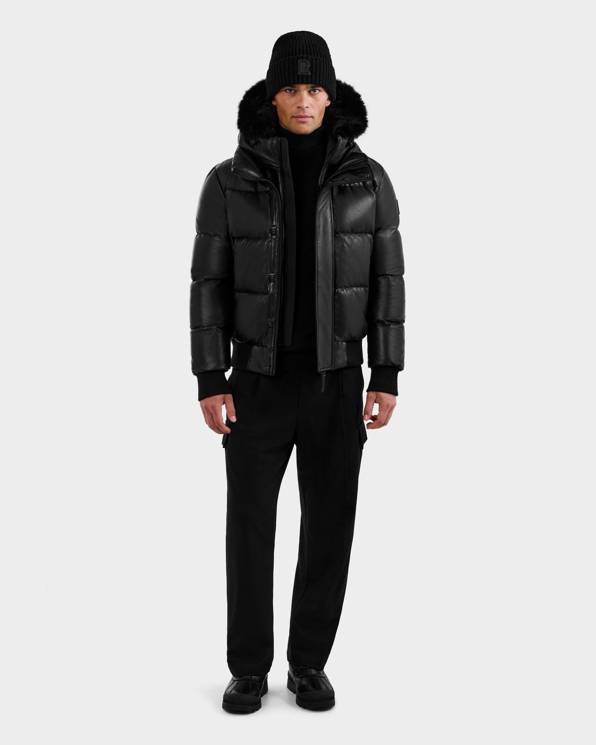 A man faces forward against a white background wearing the RUDSAK BOND X - 6122525 BLACK lamb leather bomber jacket with fur-trimmed hood, black pants, sweater, knit beanie, and shoes—dressed for cold-weather protection.