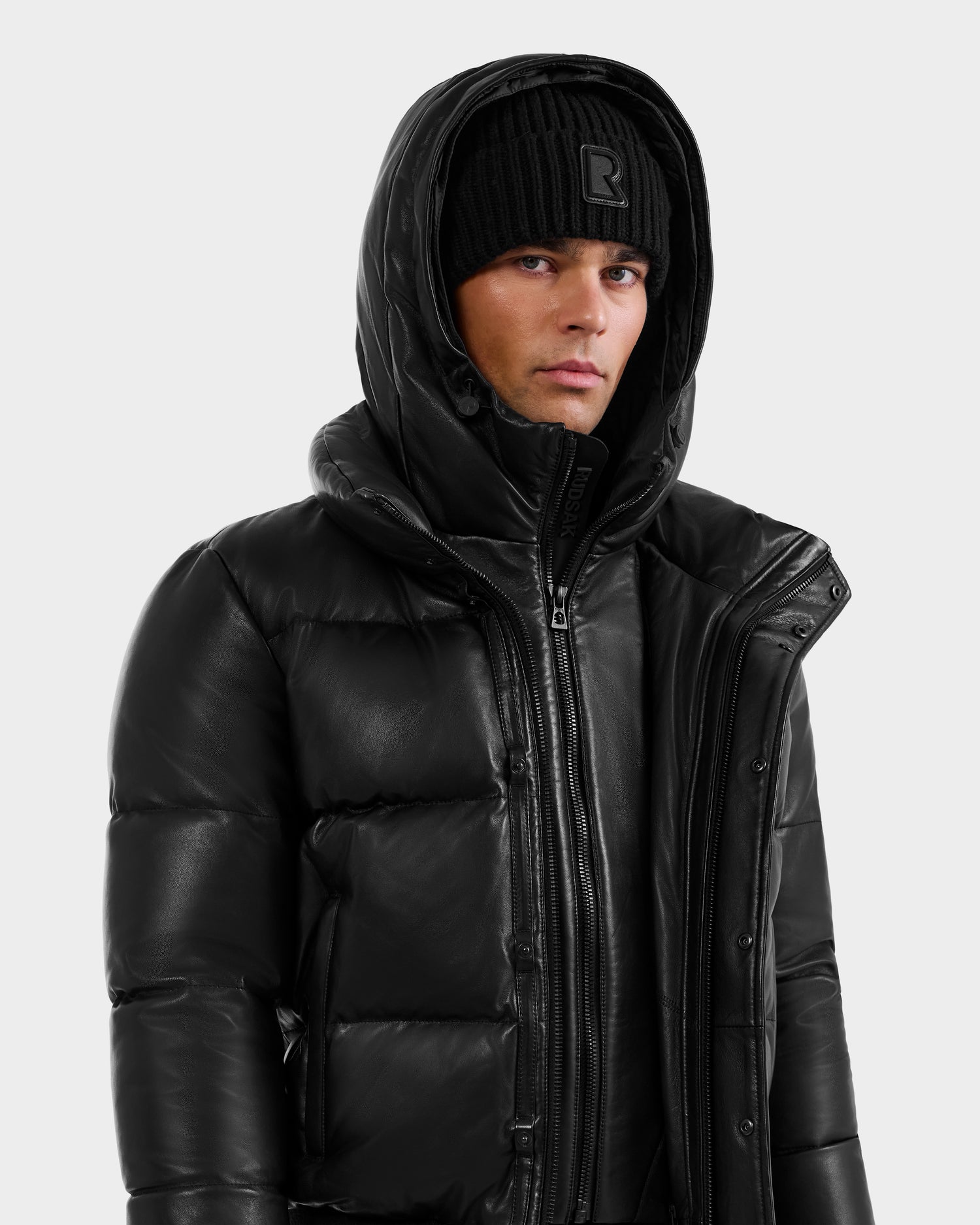 Wearing the RUDSAK BOND X - 6122525 BLACK men's down jacket with a hood and a black knit beanie, the person looks slightly to the side against a plain light background, highlighting modern cold-weather style.