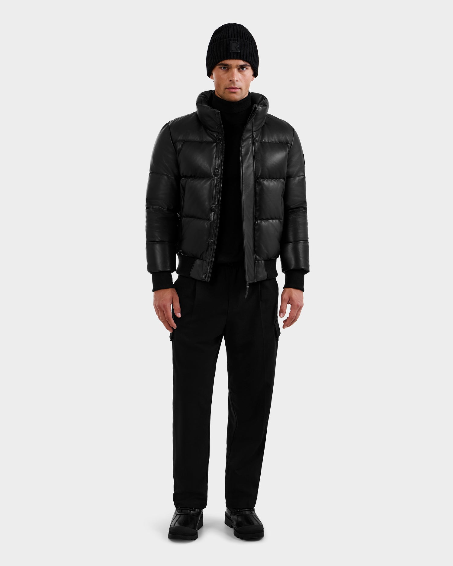 A person faces forward against a plain light gray background, wearing the RUDSAK BOND X - 6122525 BLACK men's down jacket for cold-weather protection, paired with a black beanie, turtleneck, pants, and shoes.