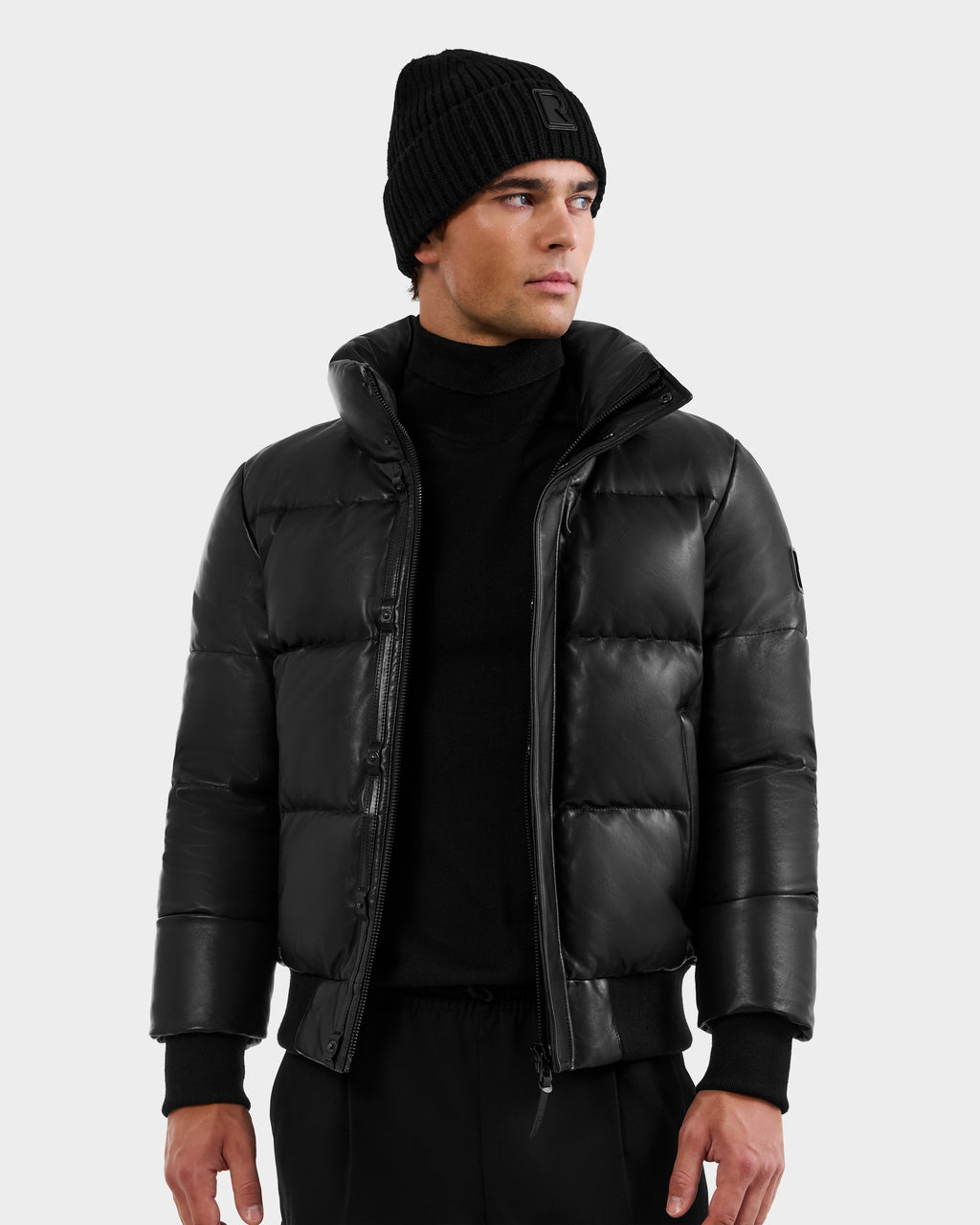 A man wearing the RUDSAK BOND X - 6122525 BLACK down jacket, black knit beanie, and black pants stands against a plain light background, looking left with a neutral expression—showcasing stylish cold-weather protection.