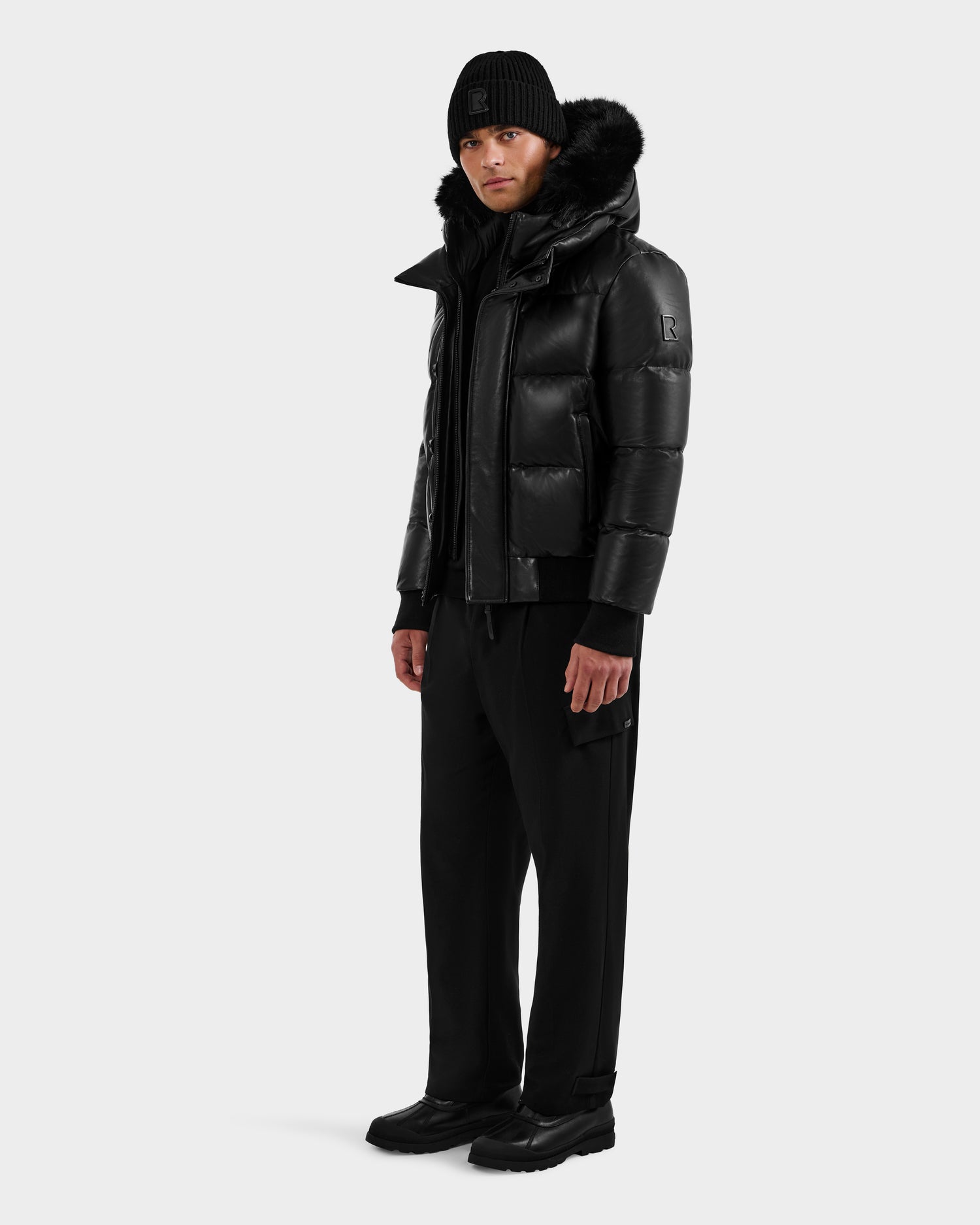A person wearing the RUDSAK BOND X - 6122525 BLACK puffer jacket with RDS-certified down and fur-lined hood, plus black beanie, pants, and shoes, stands sideways against a light gray background ready for cold weather.