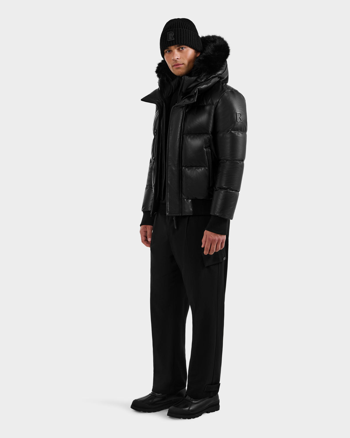 A person wearing the RUDSAK BOND X - 6122525 BLACK puffer jacket with RDS-certified down and fur-lined hood, plus black beanie, pants, and shoes, stands sideways against a light gray background ready for cold weather.