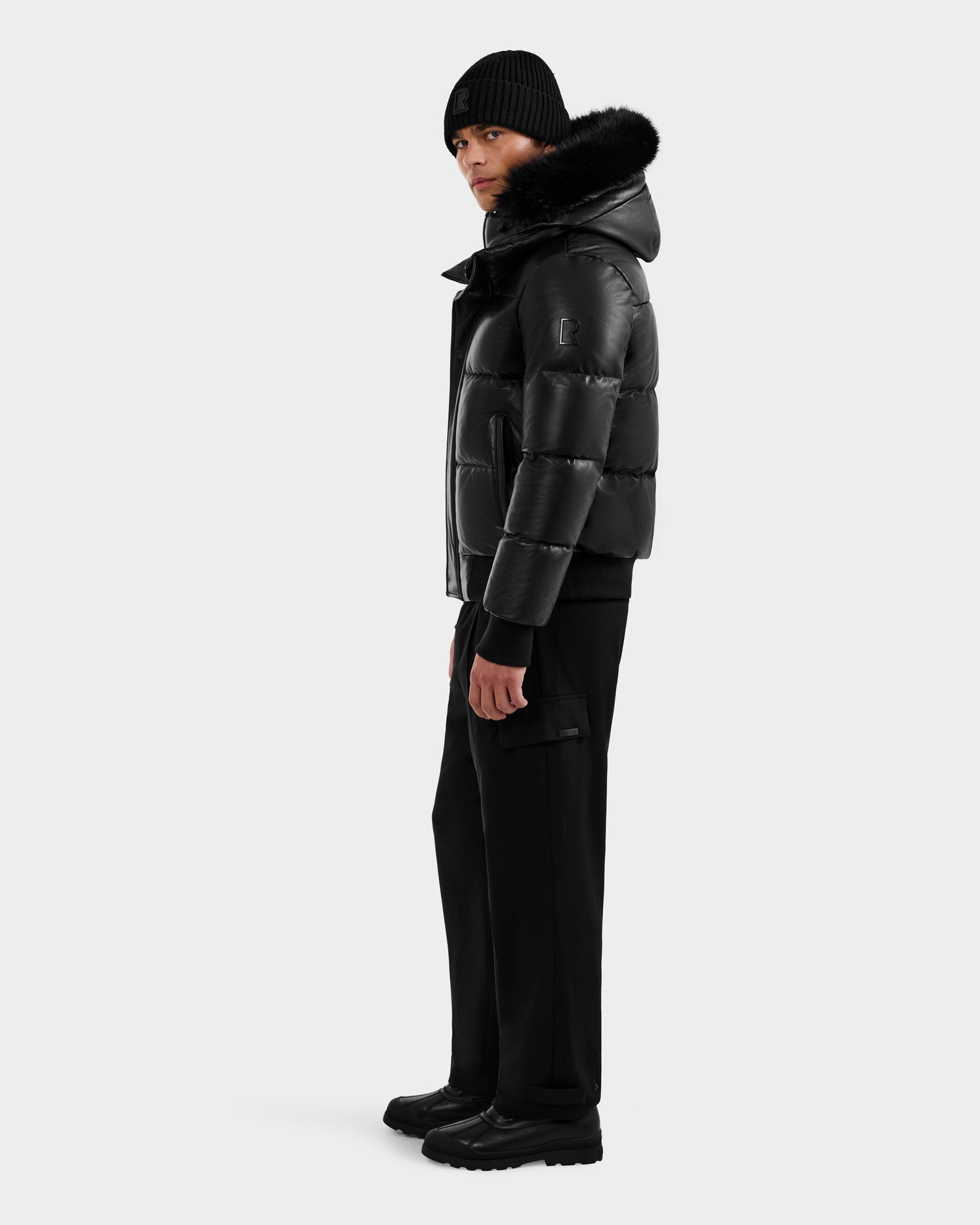 Wearing the RUDSAK BOND X - 6122525 BLACK puffer jacket with RDS-certified down and a fur-lined hood, black pants, boots, and a black beanie, a person stands slightly turned to the side against a plain backdrop.