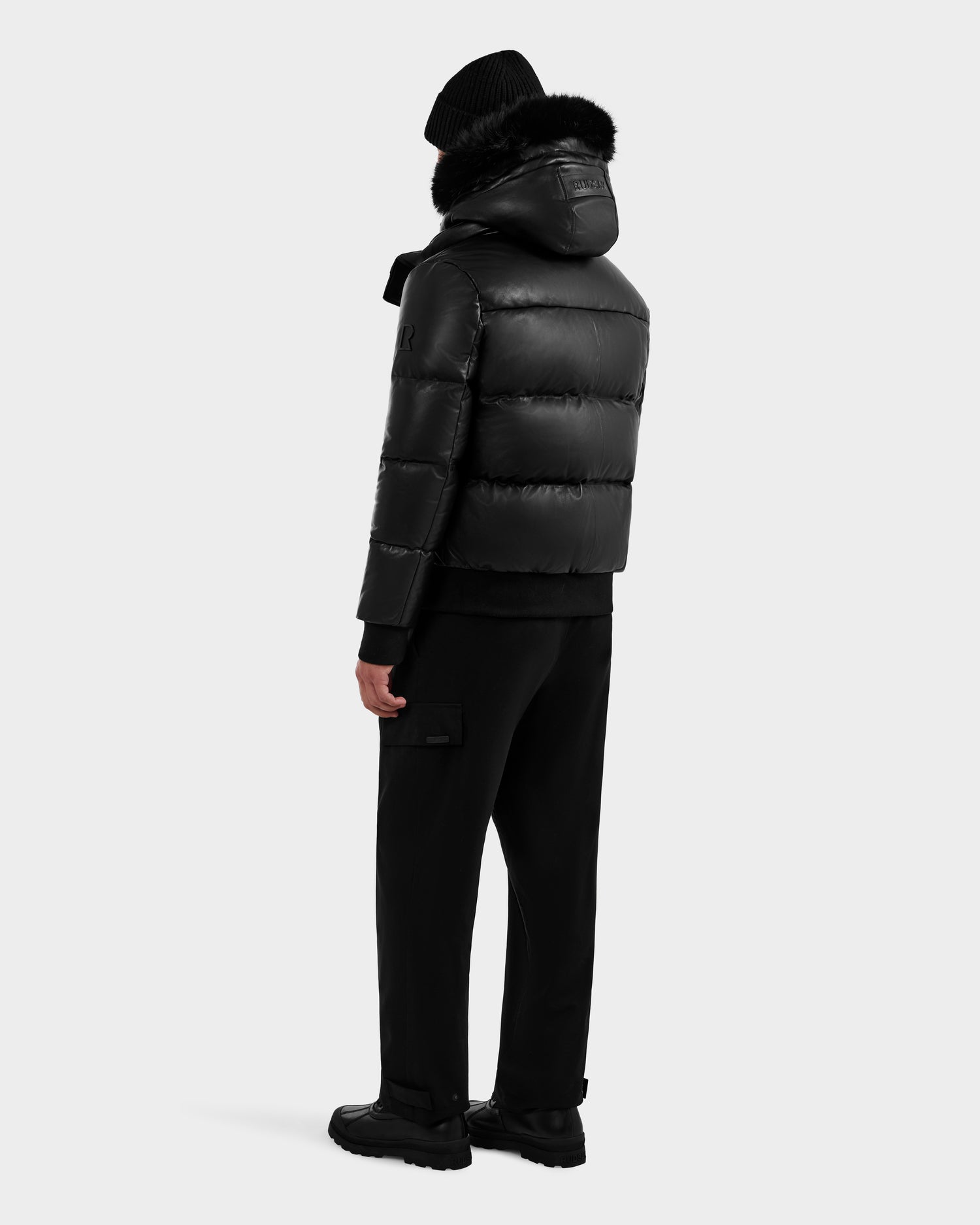 A person faces away wearing the RUDSAK BOND X - 6122525 BLACK men’s down jacket with a fur-trimmed hood, black pants, boots, and beanie against a light gray background—ideal cold-weather protection.