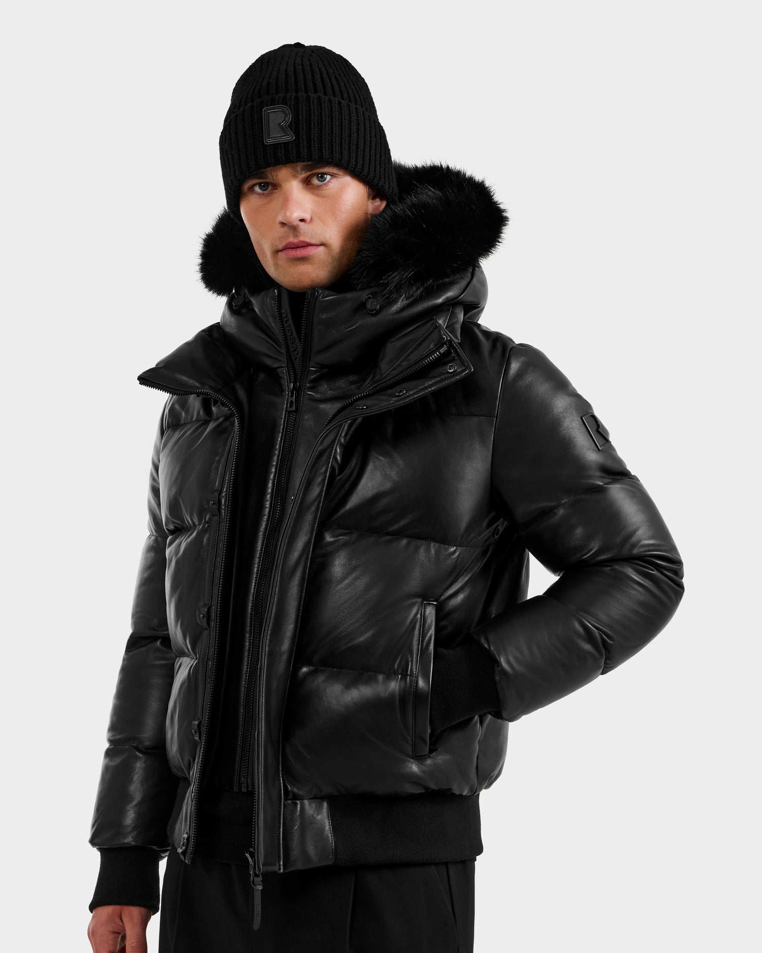 Wearing the RUDSAK BOND X - 6122525 BLACK puffer jacket with RDS-certified down and a fur-lined hood, plus a black knit beanie, this person stands against a plain light background—perfect for superior cold-weather protection.