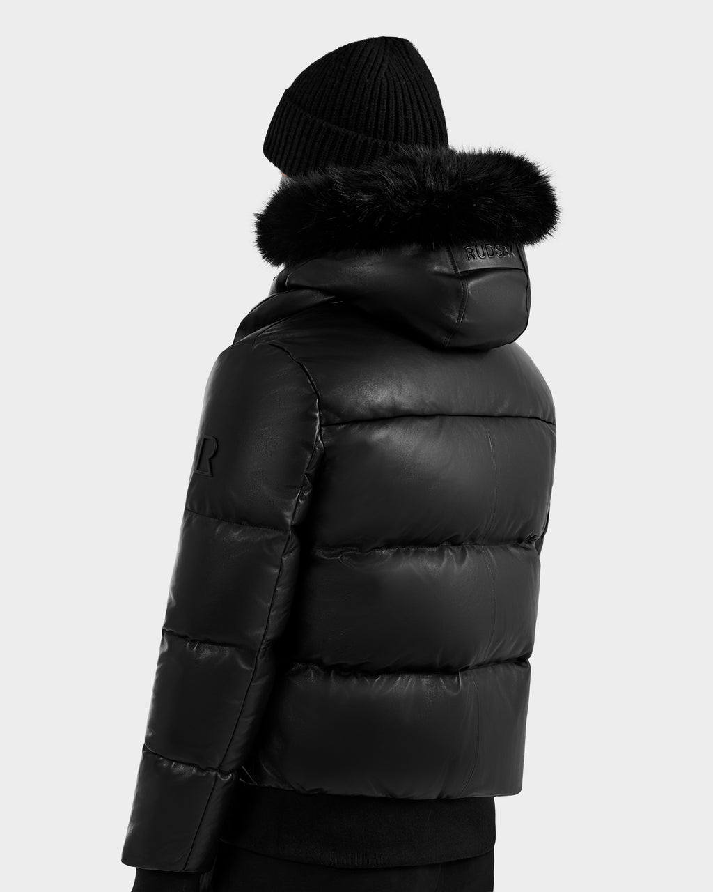 A person is shown from the back wearing RUDSAK's BOND X - 6122525 BLACK puffer jacket with RDS-certified down and a fur-lined hood, plus a black knit beanie, set against a plain light background.