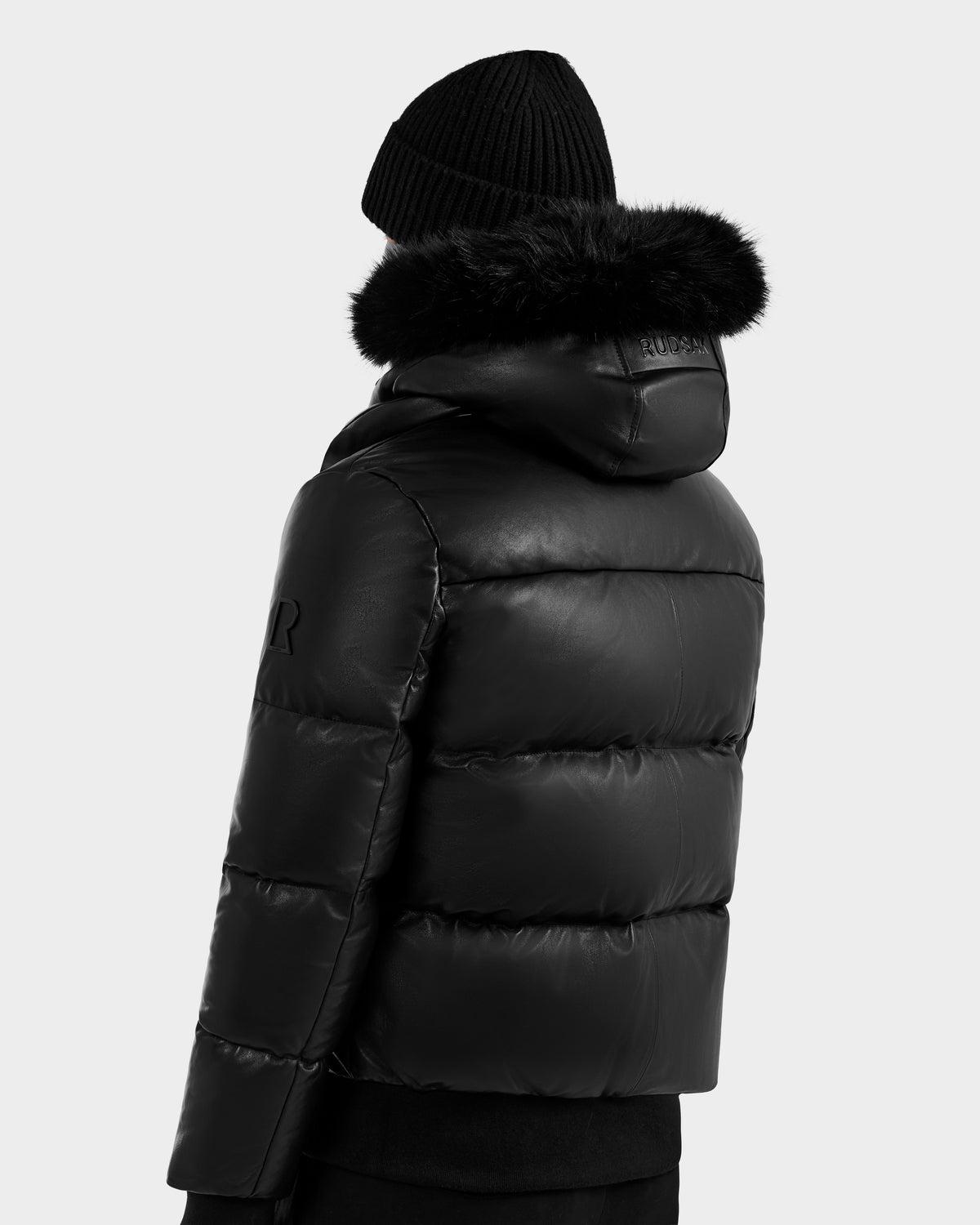 A person is shown from the back wearing RUDSAK's BOND X - 6122525 BLACK puffer jacket with RDS-certified down and a fur-lined hood, plus a black knit beanie, set against a plain light background.