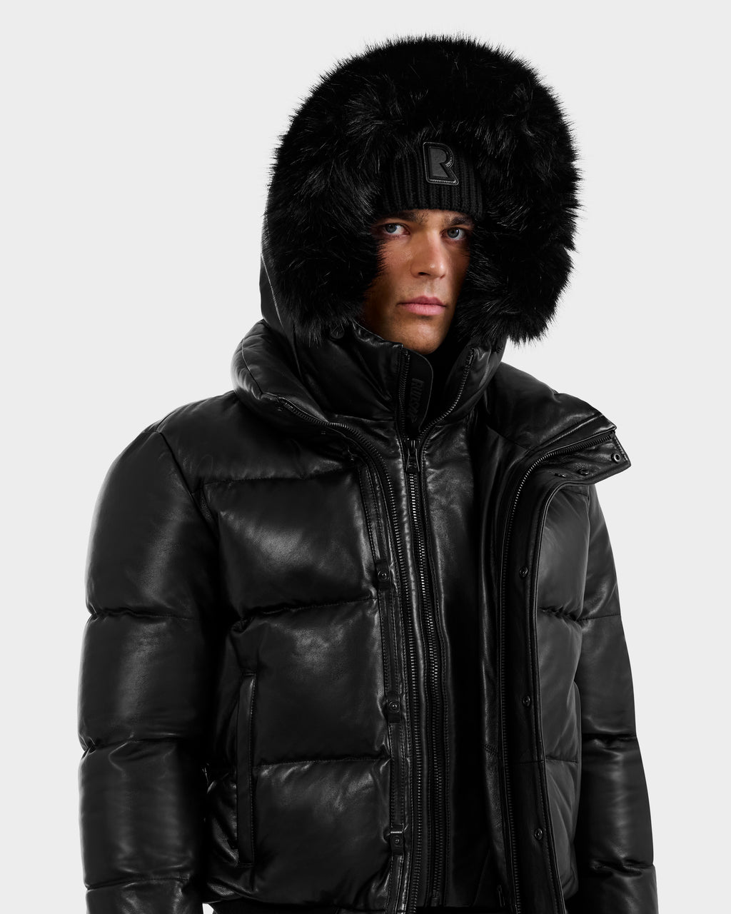 A person wears the RUDSAK BOND X - 6122525 BLACK puffer jacket with RDS-certified down and a faux fur-lined hood, paired with a black knit beanie, standing against a plain white background and looking slightly to the side.