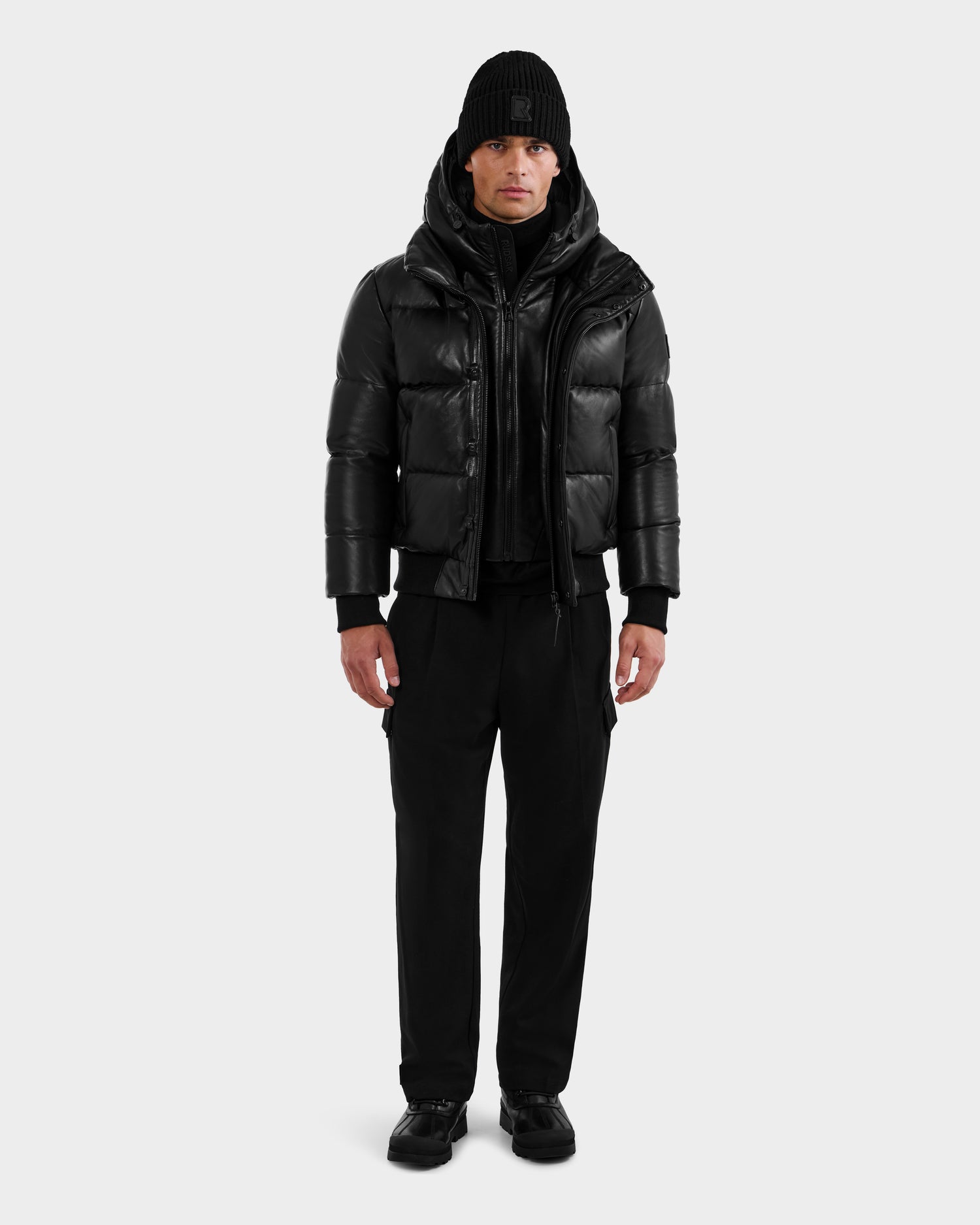 A man stands facing forward in a RUDSAK BOND X - 6122525 BLACK men’s down jacket, black beanie, pants, and shoes against a light gray background—styled for cold-weather protection.